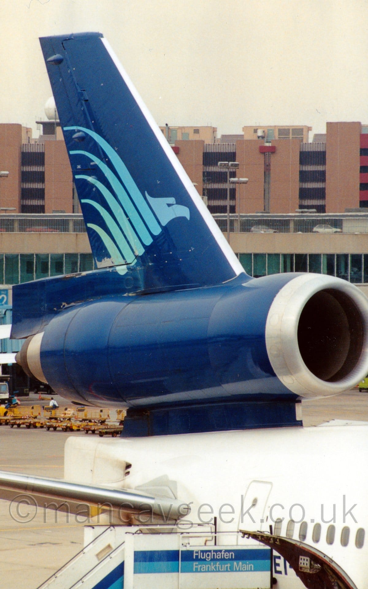 Close-up of the dark blue tail and centre engine of a white, 3 engined jet airliner parked facing to the right. There is a stylised, light blue flying bird with wings stretched out on the tail. A set of mobile airstairs is attached to the closed rear door under the tail, with a pair of dark and light blue stripes with white writing "Flughafen Frankfurt Main"  on the side. A door in the loser fuselage is hinged to open upwards, partially obscuring the planes registration marks, so only "EI-" is visible. In the background, Brown and grey buildings stretch across the frame, under grey skies.