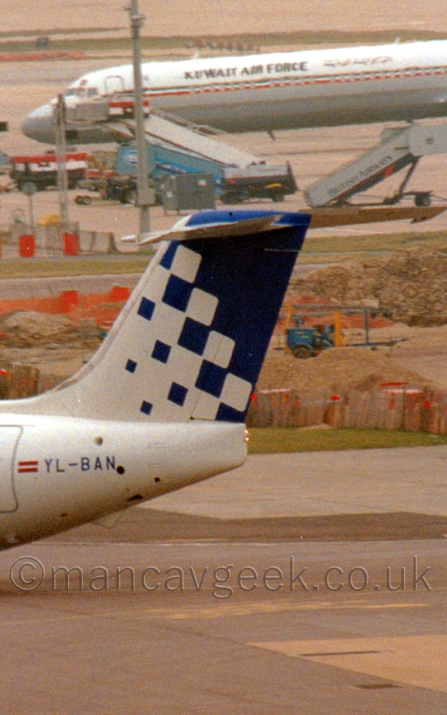 Close-up of the white and blue tail of a white jet airliner taxiing from right to left. The plane's tail starts as plain white on the left, and a series of dark blue blocks start appearing, growing larger and more numerous as they move to the right, until the fill the back end of the tail. The registration "YL-BAN" is on the rear fuselage, just aft of the cabin rear door and a small Latvian flag ( thick red bars on the top and bottom, with a thinner white bar across the middle.) Concrete apron fills the foreground, with a small building site behind the planes tail, and the forward fuselage of a white and grey jet airliner with the words "Kuwait Air Force" on the side further back.