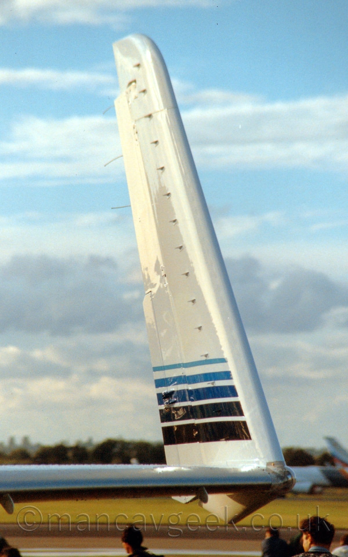 Close-up of the inside face of the left upturned wingtip of a white BizProp parked facing to the right. There are 5 blue stripes running from front to back, pale blue and thin at the top, getting darker and thinner as they get lower, the lowest one being almost black. The heads of people walking around in the background can be seen at the bottom of the frame. A couple of out-of-focus planes can be seen in the distance, with trees behind them stretching across the lower part of the frame, the rest being filled with streaks of pale blue sky and clouds.