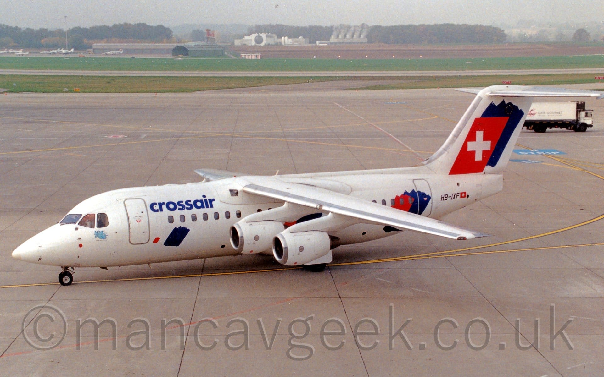 Side view of a white, high-winged, 4 engined jet airliner taxiing from right to left, following a yellow line painted on the concrete floor. There are blue "CrossAir" titles on the upper forward fuselage, and a small image of a blue flying elephant under the cockpit windows. There is a blue splotch on the lower forward fuselage, with another blue splotch overlaid with a red splotch on the rear fuselage, and again on the tail, the red splotch here being squared off and having a large white plus sign in the middle. Concrete apron fills most of the rest of the frame, with grass further back leading up to a grey hangar on the left with planes parked in front, and a rather distinctive large white building in the middle, with trees beyond, under grim grey skies.