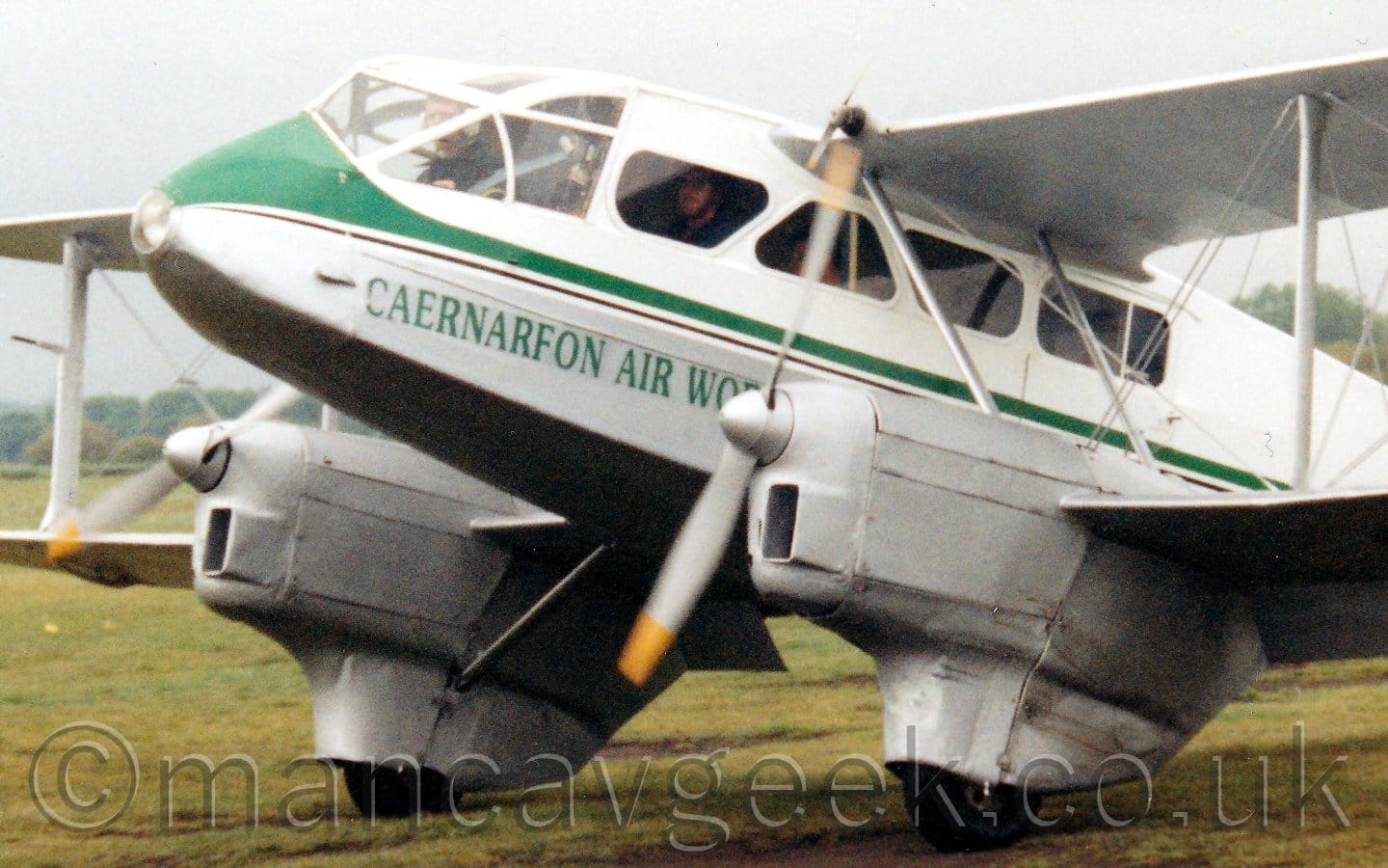 Close up of the forward fuselage of a white, twin engined bi-plane airliner taxiing from right to left on grass. The plane has a grey belly and engines, with a green stripe running along the body and green "Caernarfon Air World" titles on the lower forward fuselage. The pilot is visible in the cockpit, with passengers sitting in the cabin, visible through the triangular windows. Grey sky fills the rest of the frame.