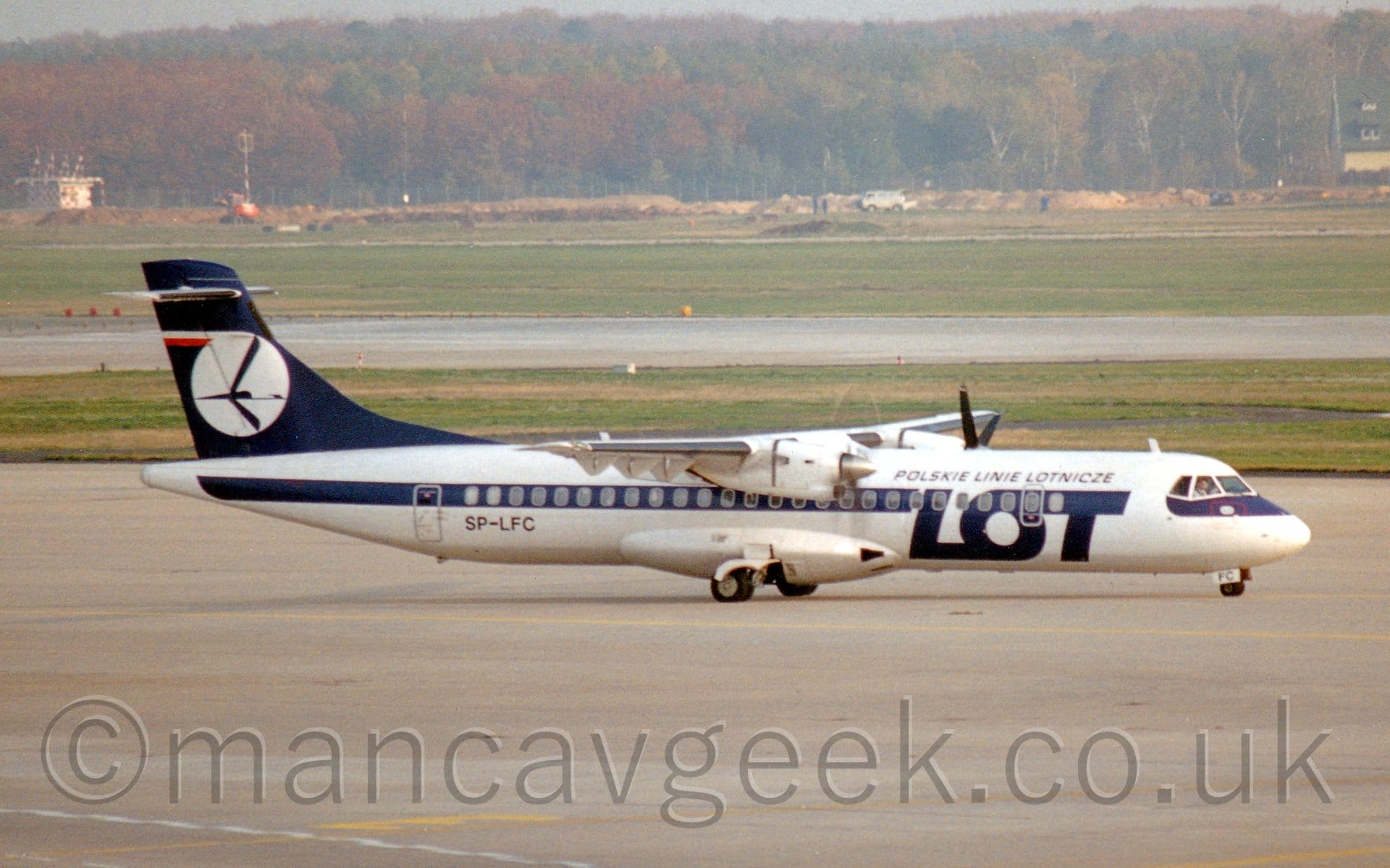 Side view of a white, high-winged, twin propellor-engined airliner taxiing from left to right. There is a thick blue stripe running along the body, covering the passenger cabin windows, with large blue "LOT" titles on the forward fuselage, and smaller "Polskie Linie Lotnicze" above that logo. The tail is the same dark blue, with a white circle containing a stylised blue flying bird. Concrete apron fills the foreground, with large areas of grass either side of a grey runway leading up to trees in the distance, under hazy grey sky.