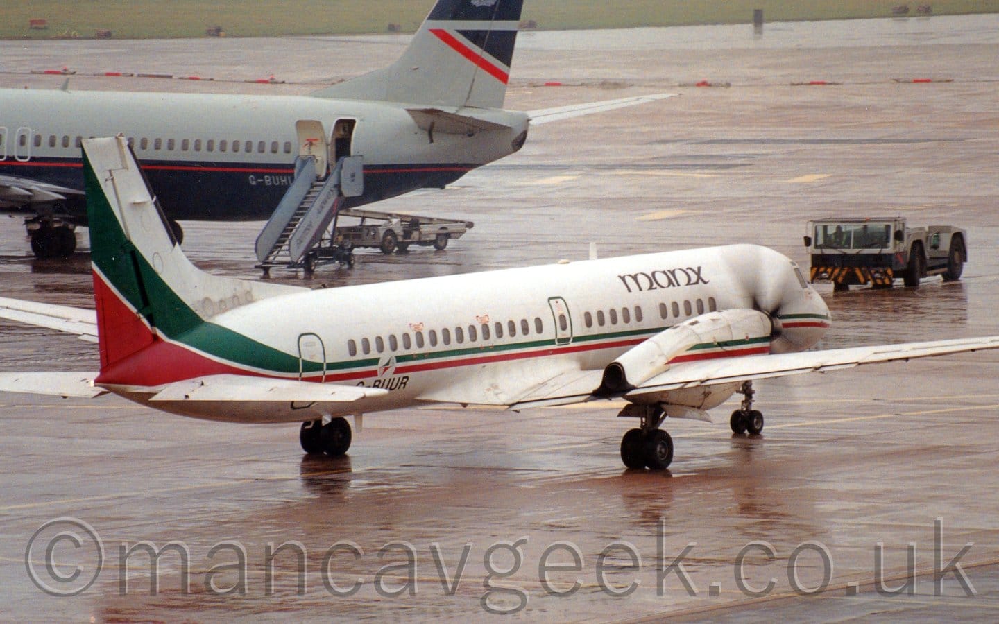 Side view of a white, twin propellor-engined airliner taxiing from left to right and slightly away from the camera, it's spinning so fast they are smeared into a blur. There is a green and red stripe running along the body just below the passenger cabin windows, getting wider as it sweeps up into the lower part of the tail. There are black "Manx" titles on the upper forward fuselage, and the registration "G-BUUR" on the lower rear, also in black. Damp, grey concrete apron fills the foreground, with more in the background, as well as grey and blue rear fuselage of a jet airliner at the top left of the frame.