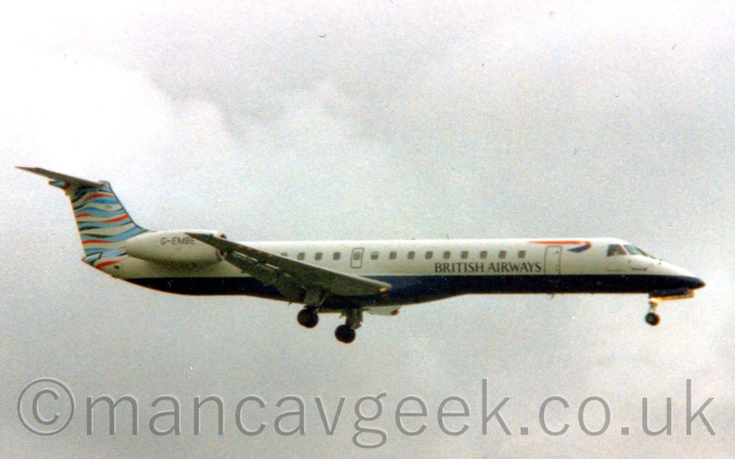 Side view of a white, twin engined jet airliner flying from left to right at a low altitude, with undercarriage lowered and flaps extended from the rear of the wings, suggesting it is about to land. The plane has a dark blue belly, with black "British Airways" on the lower forward fuselage, below a twisted red and blue ribbon. The tail has wavy light blue, dark blue, red, and white  stripes. Cloudy grey sky fills the rest of the frame.