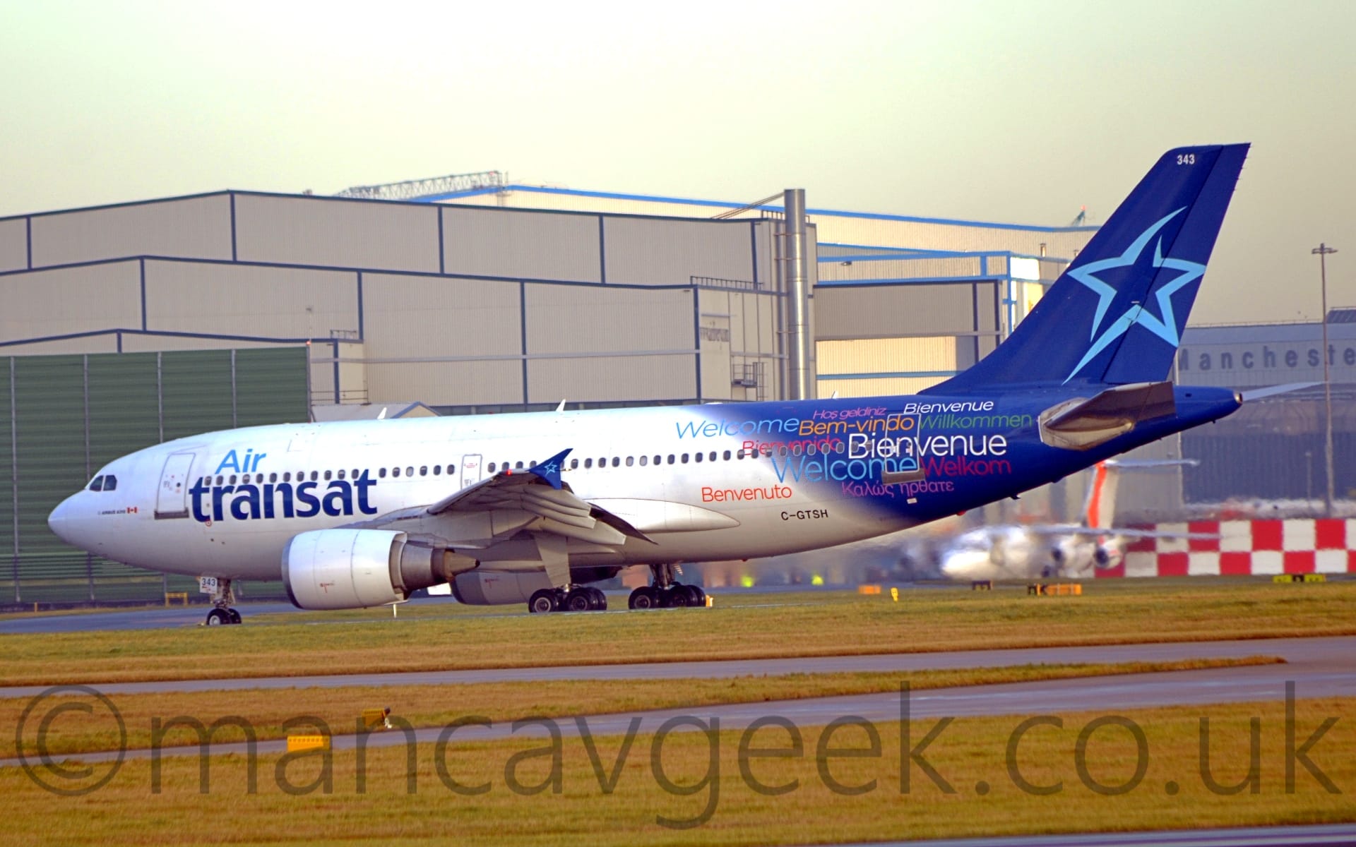 Side view of a white, twin engined jet airliner taxiing from right to left. There are light and dark blue "Air Transat" titles on the forward fuselage. The rear fuselage and tail is dark blue, with the outline of a light-blue star on the tail, and variations of the phrase "Welcome" in various languages ("Bienvenue", "Benvenuto", "Welkom", Wilkommen", and more) in various colours covering the rear fuselage. Large areas of green grass split by taxiways fill the foreground, with grey hangars dominating the background, under grey skies.