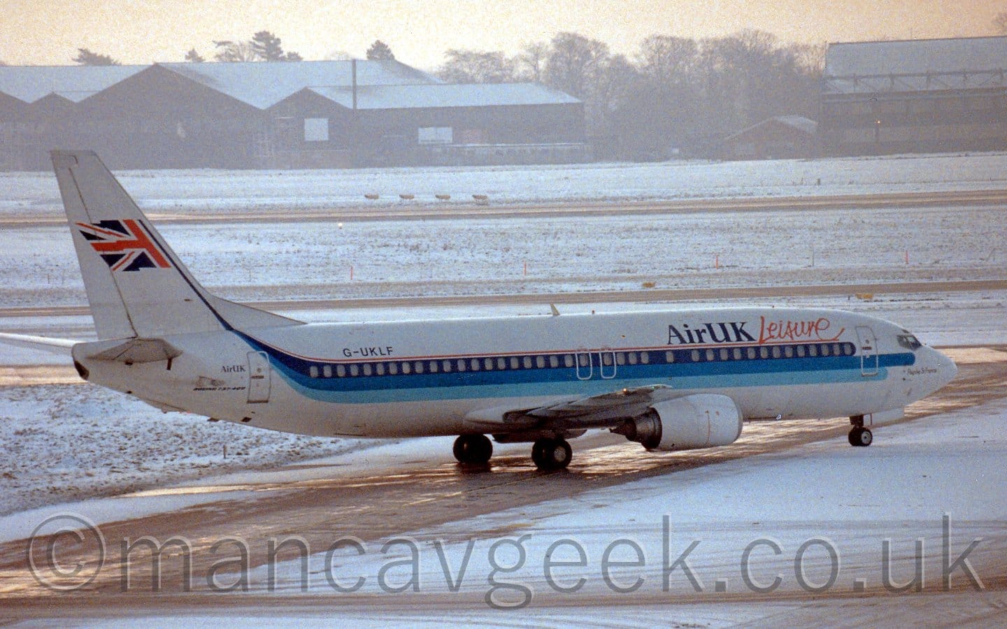 Side view of a white, twin engined jet airliner taxiing from right to left. The plane has a light blue, mid blue, and dark blue stripe running along the body, getting narrower as it sweeps up towards the tail, where it acts almost as a flag pole for a waving half UK flag. There are dark blue "Air UK" titles on the upper forward fuselage, with seemingly hand-written "Leisure" titles next to it. The registration "G-UKLF" is on the upper rear fuselage, in black. There are small "Air UK" titles just aft of the rear door, also in black. A light layer of snow fills most of the foreground, only the taxiway the plane is on is clear, shining damply in the early morning light. More snow covers grass in the background, with more taxiways and a runway clear, leading up to snow-covered hangars in the distance, under bright but hazy skies.