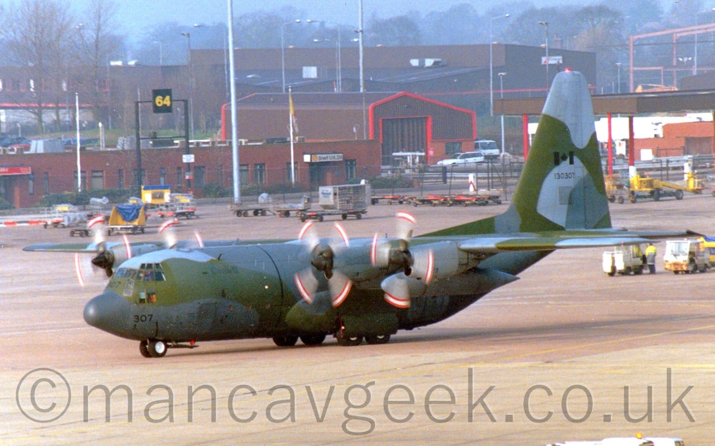 Side view of a high-winged, 4 propellor-engined military transport aircraft taxiing from right to left. The plane is covered in a green and grey camouflage scheme. There are faint "Canada" titles on the upper forward fuselage, and the number "307" on the nose. There is a black Canadian flag on the tail, above the number "130307". The red and white tips of the spinning propellors make distinct arcs as they turn. Grey concrete apron fills the foreground, with more apron at the rear, with parked luggage carts and other moveable airport furniture scattered around. Brown buildings are clustered together beyond that, with trees at the back under grey skies.