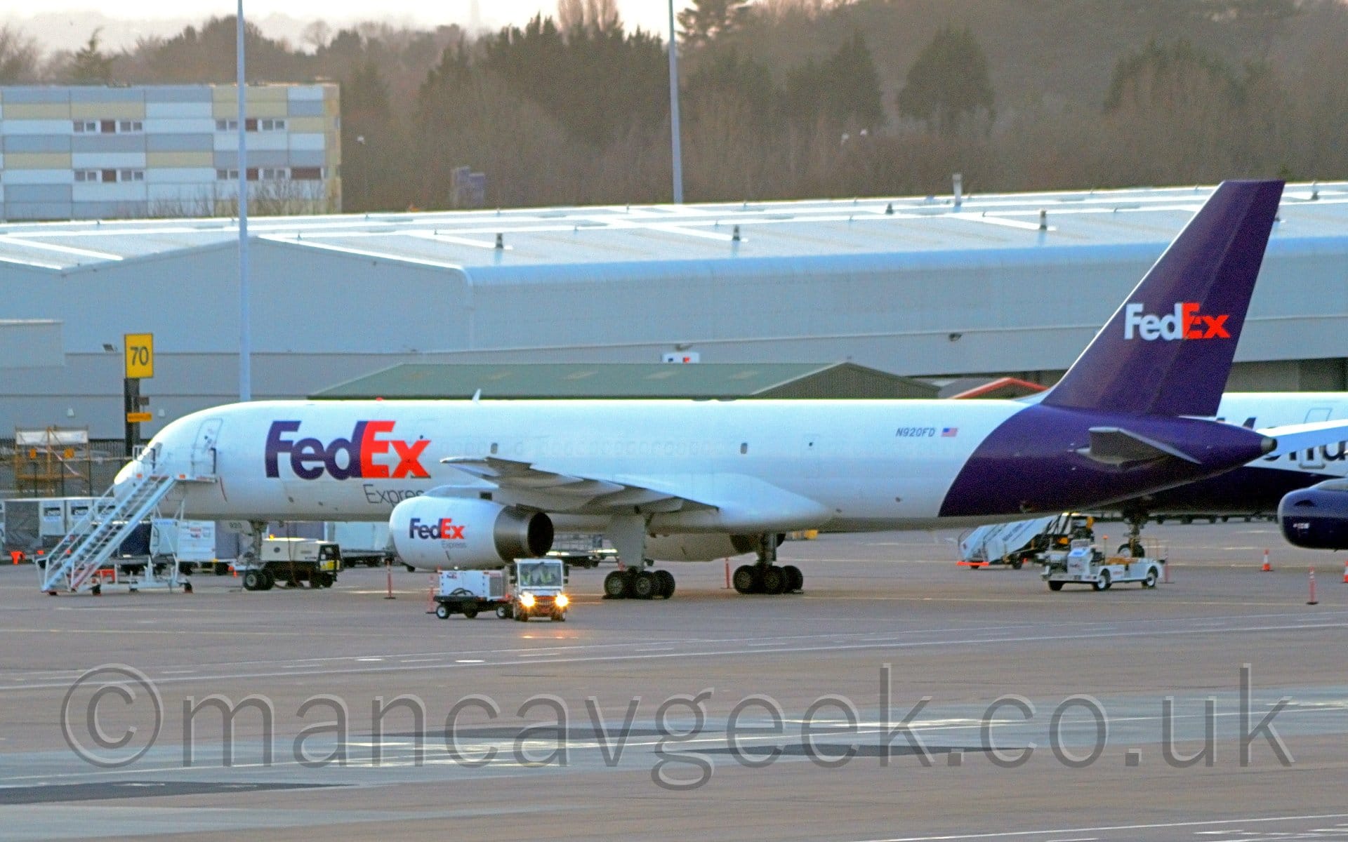 Side view of a white, twin engined jet airliner parked facing to the left and slightly away from the camera. There are purple and red "FedEX" titles on the forward fuselage and on the sides of the engine pods, repeated in white and red on the purple tail. There is a small, white, articulated truck parked next to the planes left engine, it's front turned towards the camera, lights shining down the lens. There is a a set of white mobile airstairs by the closed forward door. Another white and purple plane is parked behind this plane's tail. Wide grey buildings stretch across the frame in the background, with trees beyond that, under bright but hazy sky.