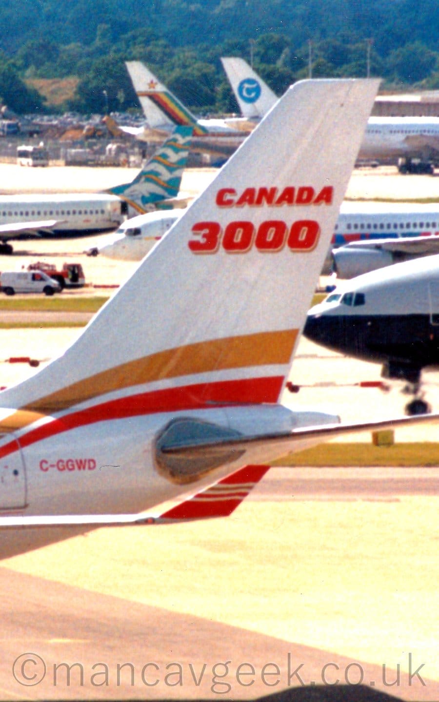 Close-up of the white rear fuselage and tail of a jet airliner  taxiing to the left. There is an amber and red stripe running up into the lower reaches of the tail from the body, as well as red "Canada 3000" titles with an amber drop-shadow.# The registration "C-GGWD" is on the upper rear fuselage in red. The plane's red upturned wingtip is visible under the tail, with white and amber stripes running along the side. In the background, the noses and tails of several colourful jet airliners fill the frame, with trees rising up a hill in the distance.