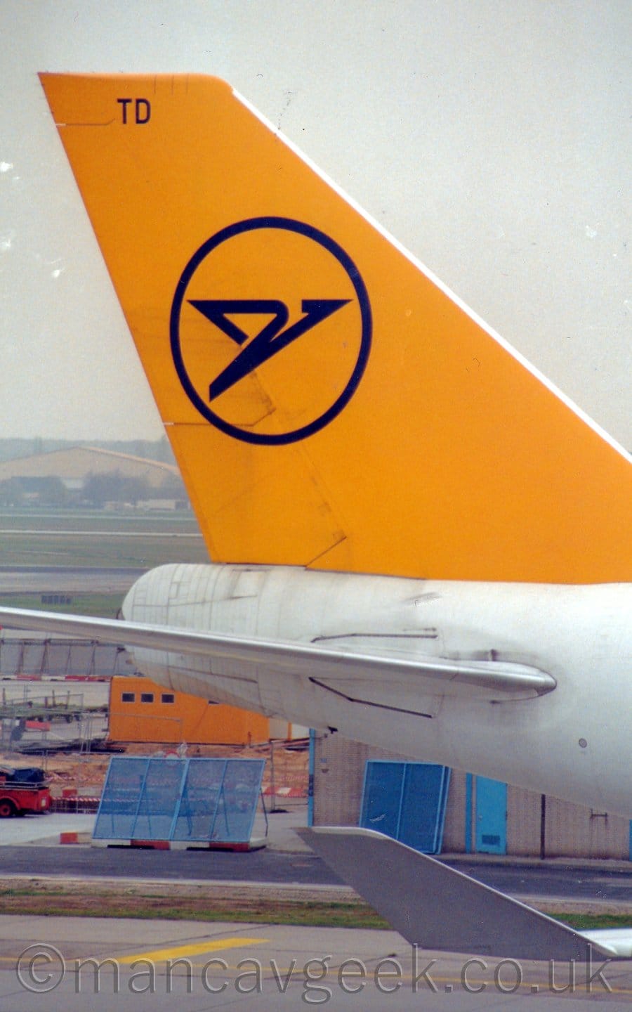 Closeup of the white rear fuselage and yellow tail of a very large jet airliner taxiing from left to right. There is the blue outline of a circle on the tail, with a stylised blue flying bird in the middle. The plane's white, upturned wingtip is visible in the bottom right corner of the framer. Blue metal fences and a yellow pre-fabricated are visible under the plane's rear fuselage. Behind the tail is a view across the airfield to a creamy-coloured hangar, under grey skies.