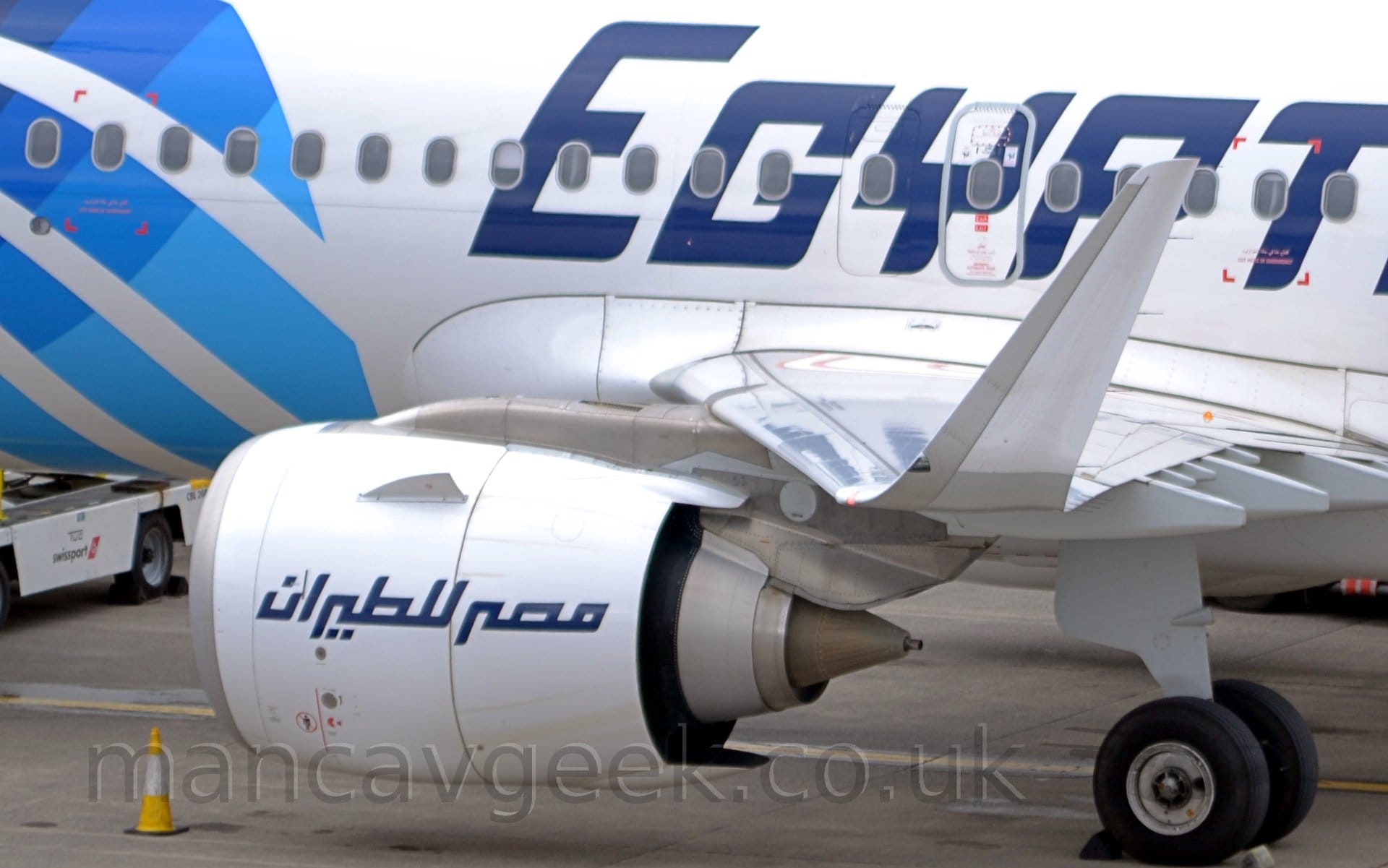 Close-up of the up-turned left wingtip and engine of a white, twin engined jet airliner parked on it's stand facing to the left. The plane has large white "EgyptAir" titles on the side of the fuselage in blue (the word "Air" being mostly out of frame), repeated in an Arabic script on the side of the engine pod. The wingtip is white, with a grey leading edge. There are diagonal light blue stripes towards the front of the fuselage, and a low-slung white truck visible under the body.