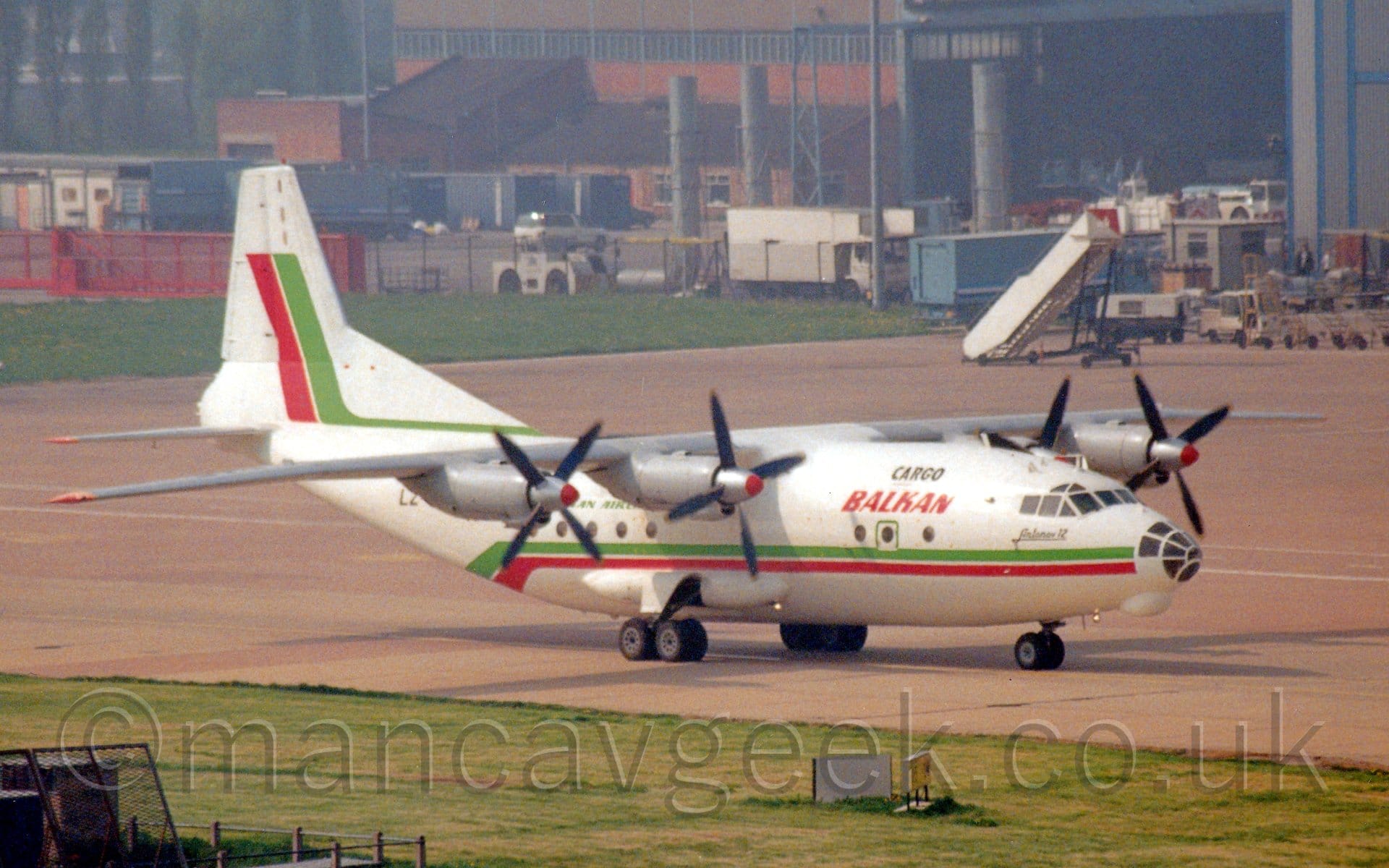 Side view of a white, high-winged, 4 propellor-engined cargo plane with a heavily glazed nose cone taxiing from left to right. A green and red stripe runs along the body, sweeping under the tail, with another, similar stripe running along the base of the tail before sweeping up at the rear. There are red "Balkan" titles on the upper forward fuselage, under smaller black "Cargo" titles. Green grass fills the foreground, with grey concrete apron and green grass leading up to a couple of hangars in the distance.