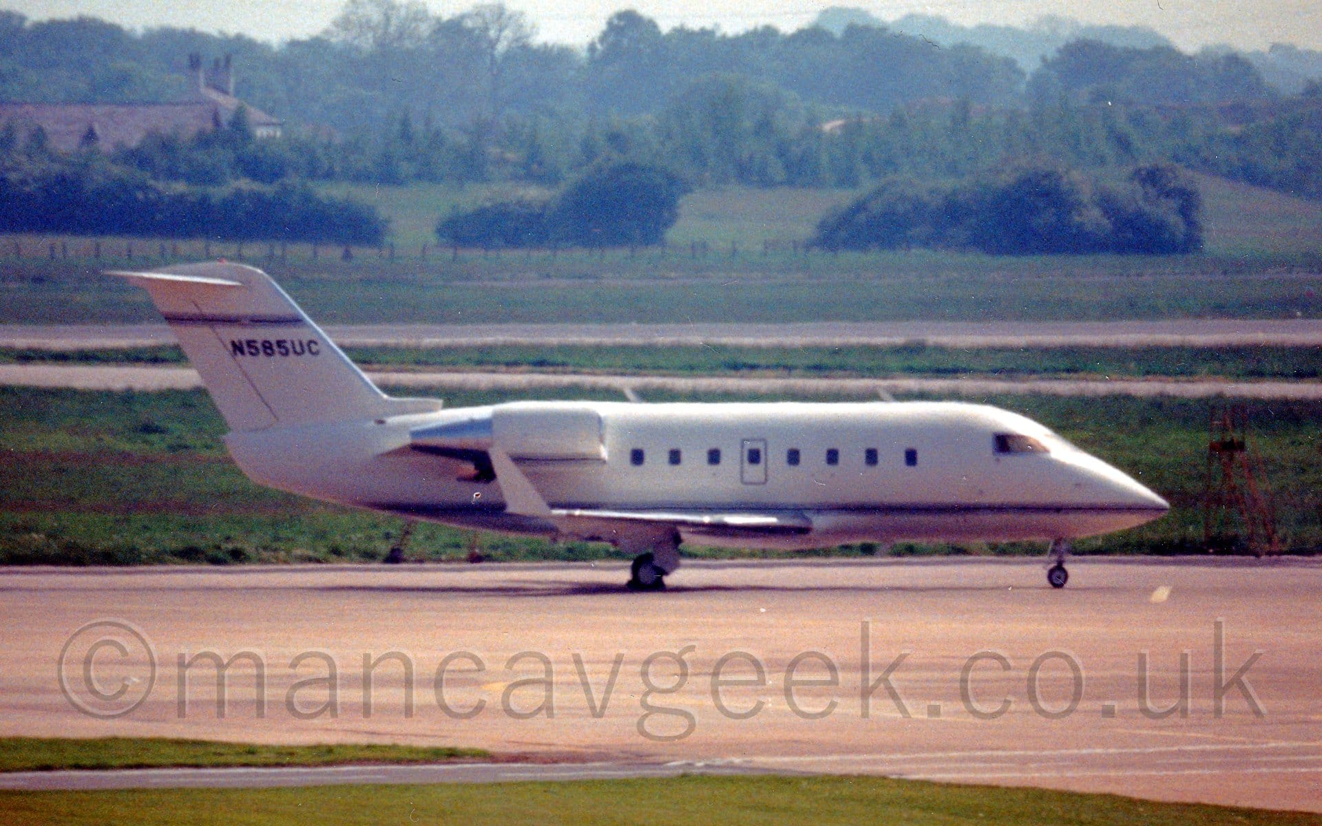 Side view of a white, twin engined BizJet parked facing to the facing to the right. The plane is almost entirely white, with a thin dark blue stripe running along the body, and the black registration "N585UC" under another thin dark blue stripe on the tail. Grey concrete apron fills most of the foreground, with grass in the background stretching off to trees in the distance, under hazy grey sky.
