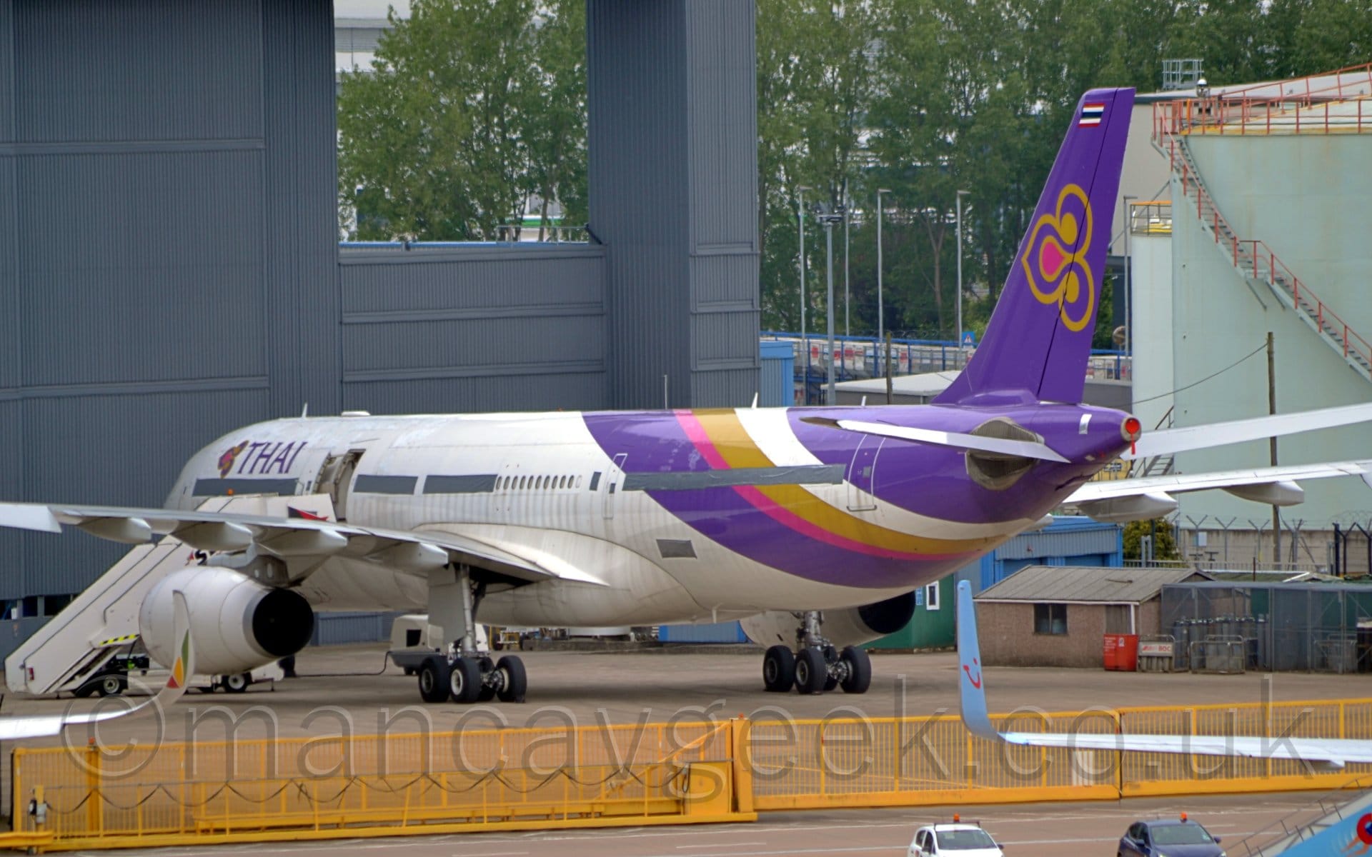Side view of a white, twin engined jet airliner parked facing to the left and slightly away from the camera. The plane is mostly white, with purple, magenta, gold, and white diagonal bands around the rear fuselage, and a purple tail with a stylised gold and purple lotus flower in the middle. There are purple "Thai" titles next to a smaller version of that lotus logo on the upper forward fuselage. There is a set of white mobile airstairs parked by the open mid-cabin door. A yellow metal barrier stretches across the bottom of the image, with the bit on the right split down the middle so it can retract back to allow access. The wings of another couple of airliners are at the bottom of the frame, a seriously curved up-turned white wing-tip with a green, yellow, and red triangle on the outer face on the left,  and a blue, somewhat straighter up-turned wingtip with a smiley face logo coming in from the right. A large grey hangar dominates the left of the image, with a duck-egg blue fuel storage tank on the right, with trees in the background behind them.