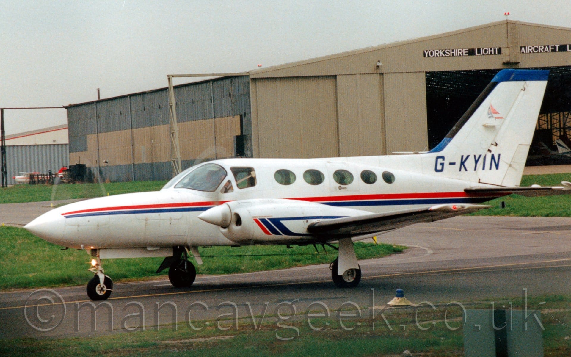 Side view of a white, twin engined BizProp taxiing from right to left. The plane has a red and blue stripe running along the body, and short, red and blue diagonal bars on the engine cowlings, the latter of which sweeps back to go over and under the wings. The registration "G-KYIN" is on the tail in blue. Green grass lines both sides of the taxiway the plane is on, with a large brown and grey hangar with the doors wide open in the background. Black boards over the hangar doors have the words "Yorkshire Light Aircraft" in large white letters. Grey sky fills the rest of the frame.