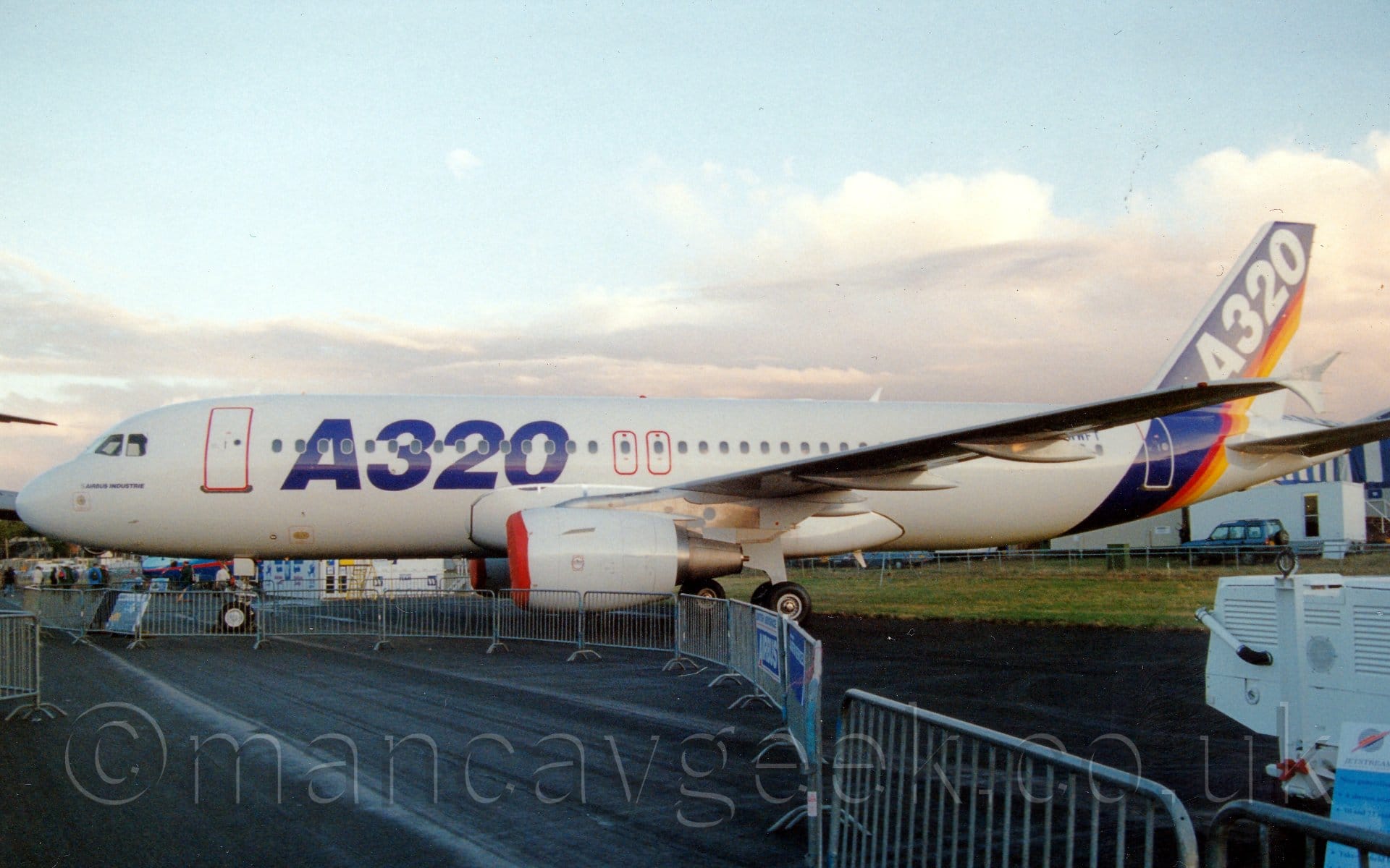 Side view of a tin-engined jet airliner parked facing to the left. The plane is mostly white, with large, blue, billboard-style "A320" titles on the forward fuselage, and a thick blue bland, with thinner red, orange, and yellow bands, wrapped around the rear fuselage and tail, with white "A320" titles on the blue section. The engine pods under the wings have red covers on the inlet. The plane is parked on a black tarmac taxiway, behind a temporary metal fence which curves through the bottom of the frame to provide a walkway for spectators. Fluffs of white cloud with hints of pale blue fill the rest of the frame.