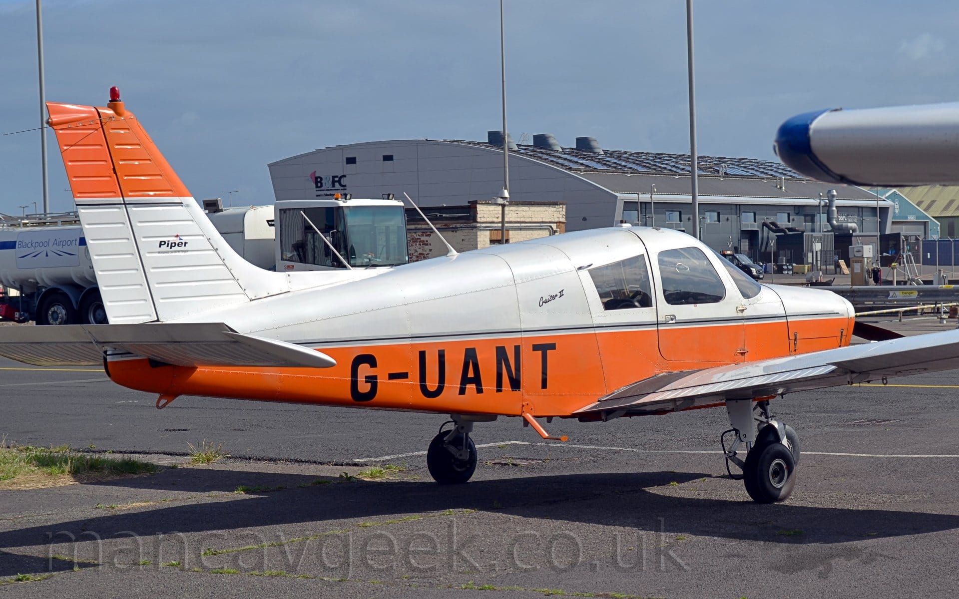 facing to the right. The plane's upper body is white, with it's lower body and tail being orange. The black registration "G-UANT" is on the lower rear fuselage. Bright sunlight casts a strong shadow on the ground under the plane. A large, grey building with a curved roof dominates the background, under grey skies.
