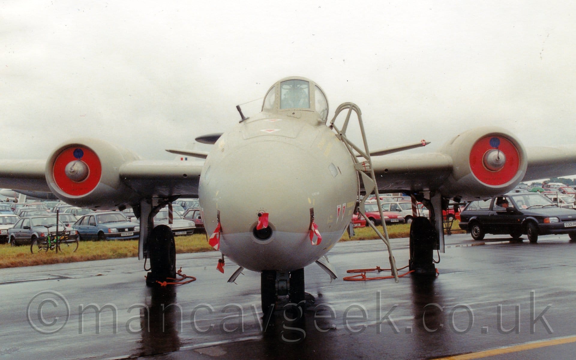 Head-on view of a twin-engined military jet bomber plane parked on black tarmac facing the camera. T plane is mostly sandy-brown, with a grey belly. The engines are buried in the wings, with red plates over the intakes, and the main landing gear is attached to the side of the engine pods. The bubble canopy over the cockpit is offset to the left of the plane, with a removeable ladder attached for the crew to access the plane. Red and white ribbons are attached to the covers of various sensors on the planes nose. Puddles on the black tarmac suggest that it has recently been raining. A field full of parked cars is in the background, stretching off into the distance, under flat grey sky.