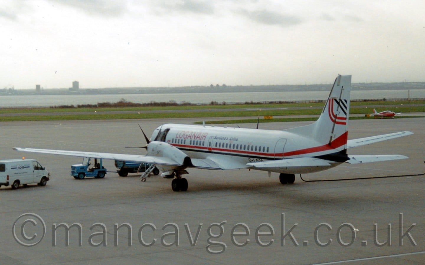 Rear view of a twin propellor-engined airliner parked facing to the left and away from the camera. The plane is mostly white, with a black and red stripe running along the body, and red "LoganAir" titles on the upper forward fuselage, with additional black "Scotlands Airline" titles on the upper rear. The forward left door is open, with a set of built-in airstairs coming down to the ground. A blue tug with an attached trailer is parked in front of the planes nose, with a white van off to the left. Grey concrete apron fills the bottom half of the screen, with green grass in the background leading up to a wide river, the other bank disappearing into grey haze.