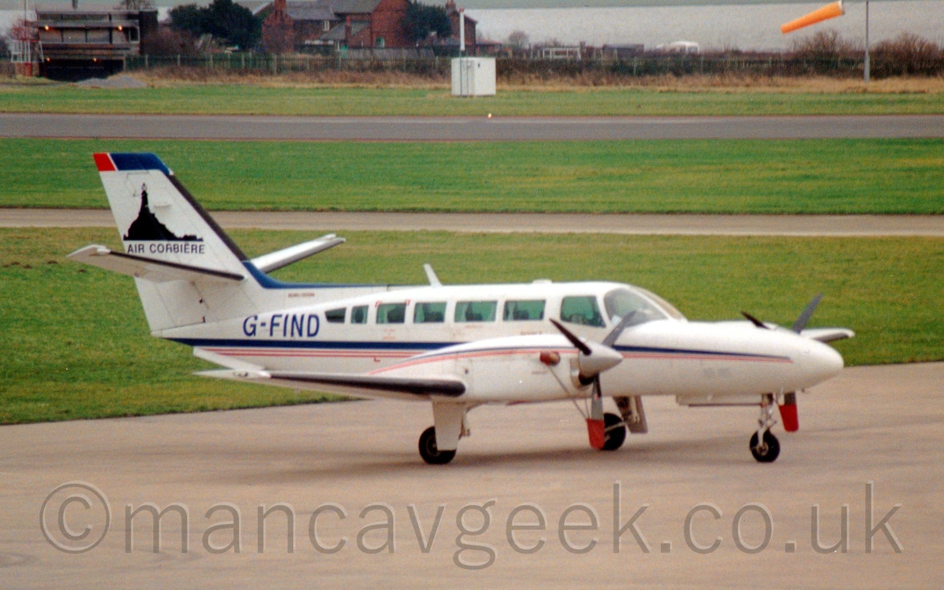 Side view of a white, twin engined BizProp parked facing to the right. There is a blue and red stripe running along the bidy, as well as over the top section of the engine cowlings over the wings. The blue registration "G-FIND" is on the upper rear fuselage. There are black "Air Corbiere" titles on the tail, below a black image of a tower on a domed hill. Grey concrete apron fills the foreground, with green grass separated by grey taxiways and a black runway leading off into the distance in the background. A red brick building stands right at the back, with a large body of water just beyond that.