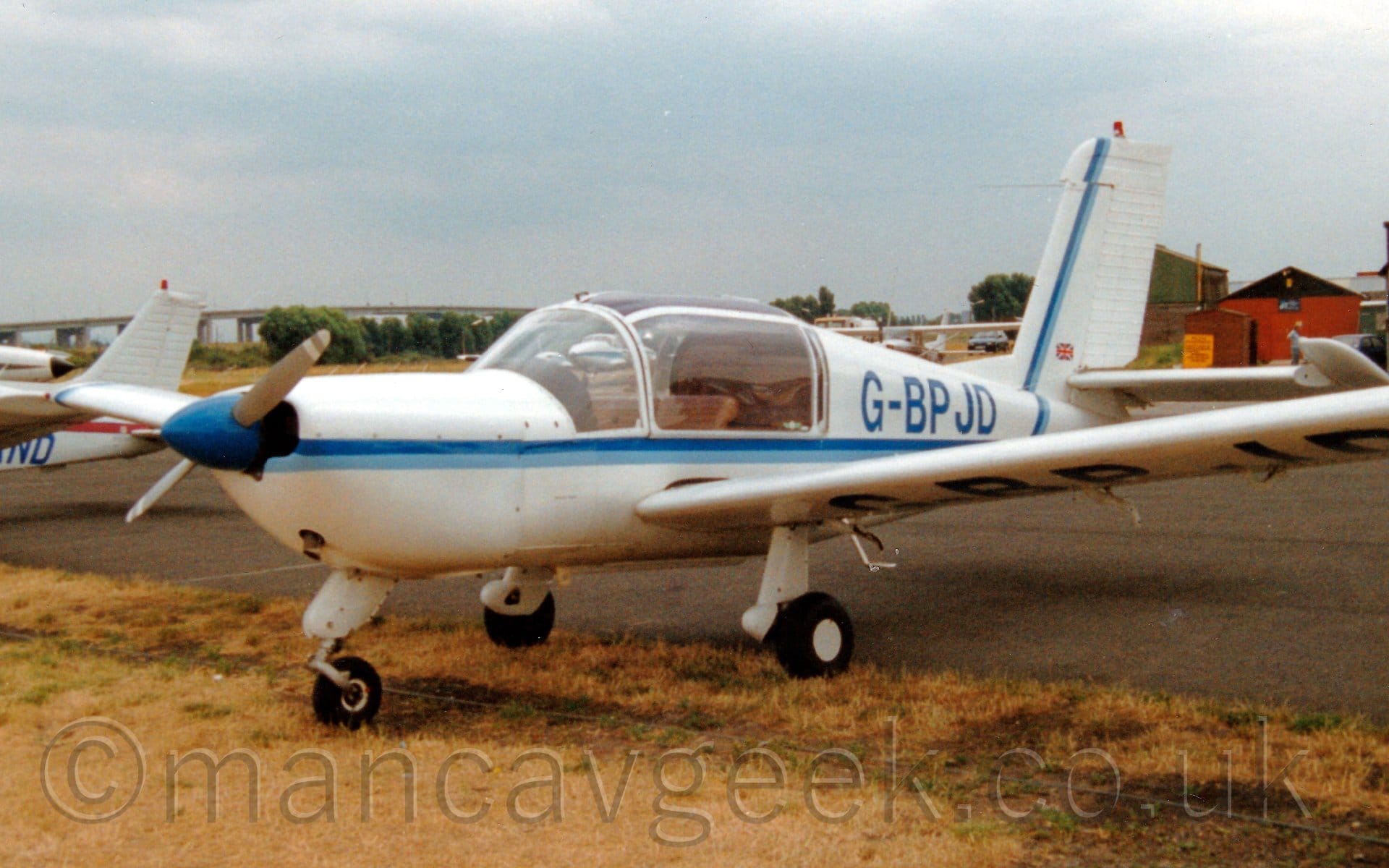 Side view of a single-engined light aircraft parked facing to the left on yellowing grass, right on the edge of a black tarmac taxiway. The plane is mostly white, with a 2-tone blue stripe running along the body before sweeping up the front of the tail. The registration "G-BPJD" is on the upper rear fuselage, as well as under the left wing. Another couple of planes are parked around it, as well as a couple of wooden buildings on the right. A long bridge reaches over the horizon on the left, with blue-grey sky filling the rest of the frame.