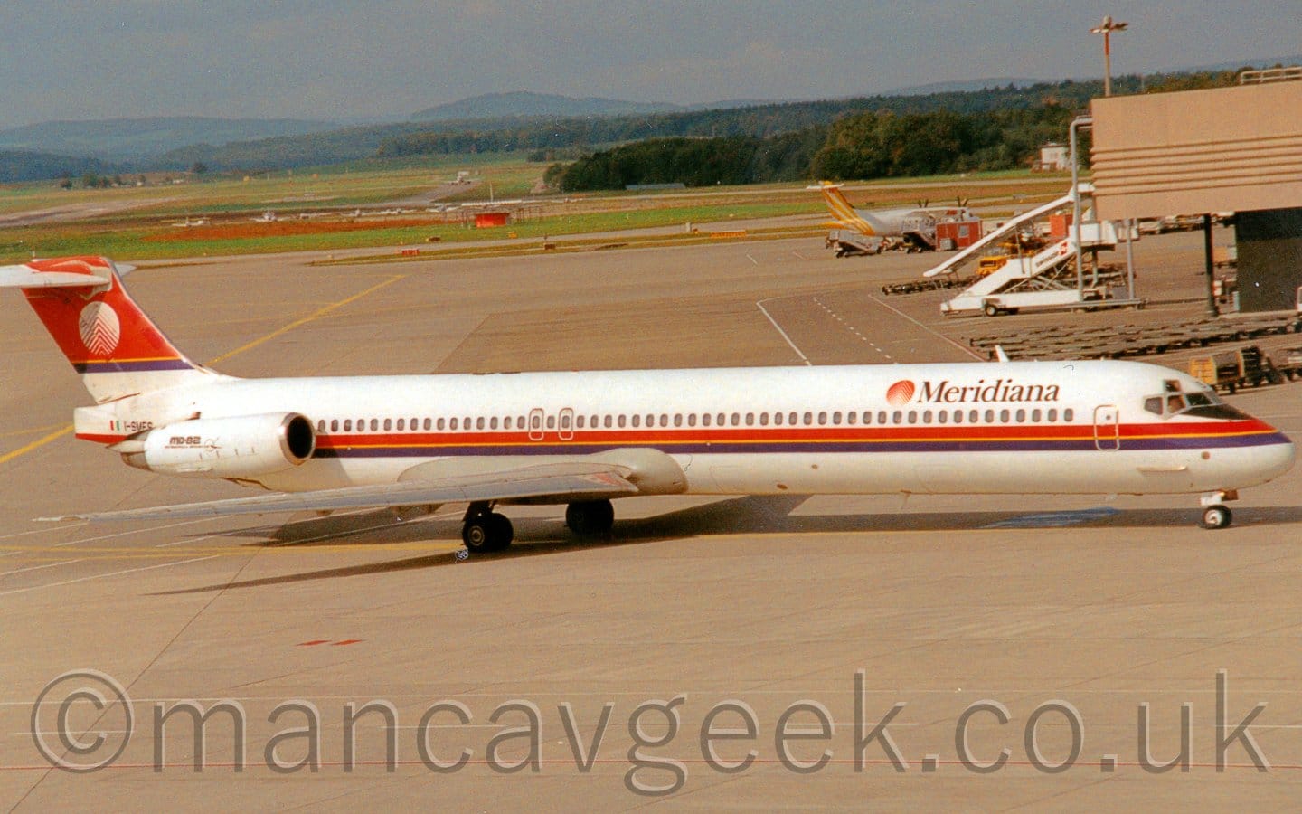 Side view of a white, twin engined jet airliner with the engines mounted on the sides of the rear fuselage, taxiing from left to right. The plane has a red and purple stripe running along the body just below the passenger cabin windows, with yellow piping down the middle. There are black "Meridiana" titles on the upper forward fuselage, next to a red and white circular logo. The tail is mostly red, with a purple stripe and yellow piping at the bottom, with the circular logo repeated, only this time with the colours inverted. The registration "I-SMES" is on the tail-cone, behind the engines, next to an Italian flag. The engines have small, black "MD-82" titles. Grey concrete apron fills the foreground, and a good chunk of the background, with a sandy-brown terminal building just edging it's way in to shot on the right. A white, high-winged, twin propellor-engined airliner with yellow stripes on the tail is parked behind a brown prefabricated hut and a couple of sets of mobile airstairs in the background. Some light aircraft are parked on grass in the distance on the left, with more grass and trees beyond that leading off to the skyline, under hazy grey sky.