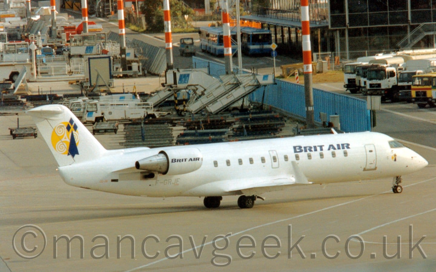 Side view of a white, twin engined jet airliner with the engines mounted on the sides of the rear fuselage, taxiing from left to right. The plane has large, dark blue "Brit Air" titles on the upper forward fuselage, repeated on the sides of the engine pods. The grey registration "F-GRJF" is on the lower rear fuselage, under the engine pod. There is a circular yellow logo overlaid with a blue vertical arrowhead with 3 blue diamonds at the top on the tail. Grey concrete apron fills the foreground, with a Aline of tall lighting poles with orange and white stripes looming over everything, reaching out of the top of the frame. A blue metal fence splits the background, on the left is a collection of white airport trucks, luggage carts, and mobile airstairs, while a gaggle of white fuel trucks are parked next to a heavily glazed building with blue busses parked outside.