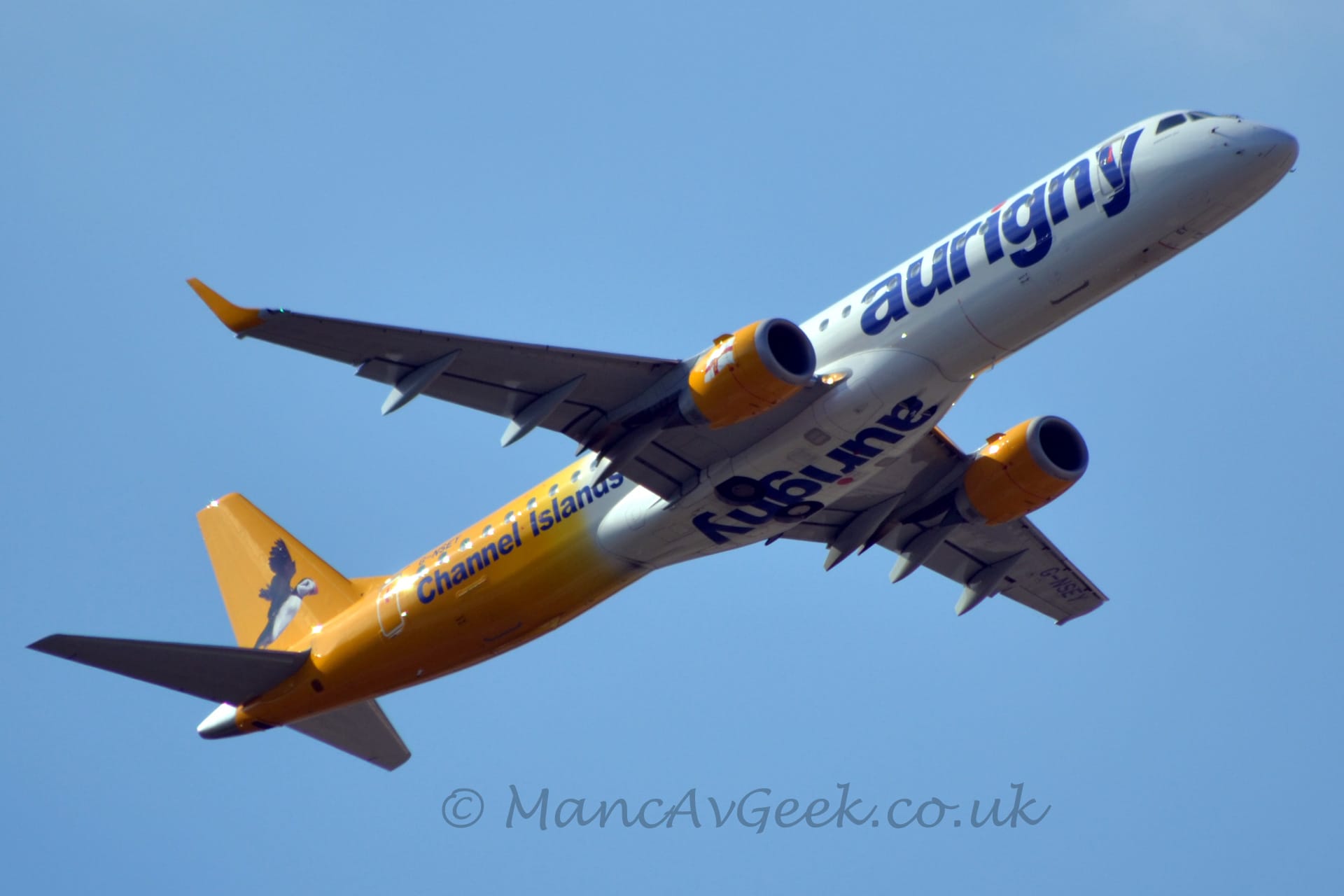 Low side view of a white and yellow, twin engined jet airliner flying from left to right at a very low altitude with flaps extended from the rear of the wings, the undercarriage safely stowed away, and the nose raised sharply, suggesting it has just taken off. The plane has a white forward fuselage, with blue billboard-style "Aurigny" titles, inverted and repeated under the belly. The rear fuselage and tail are bright yellow, with an image of a flying puffin on the tail. There are additional blue "Channel Islands" titles on the lower rear fuselage. The engine pods under the wings are the same bright yellow, with a waving white flag with a red cross in the middle, overlaid with a smaller yellow cross. Pale blue sky fills the rest of the frame.