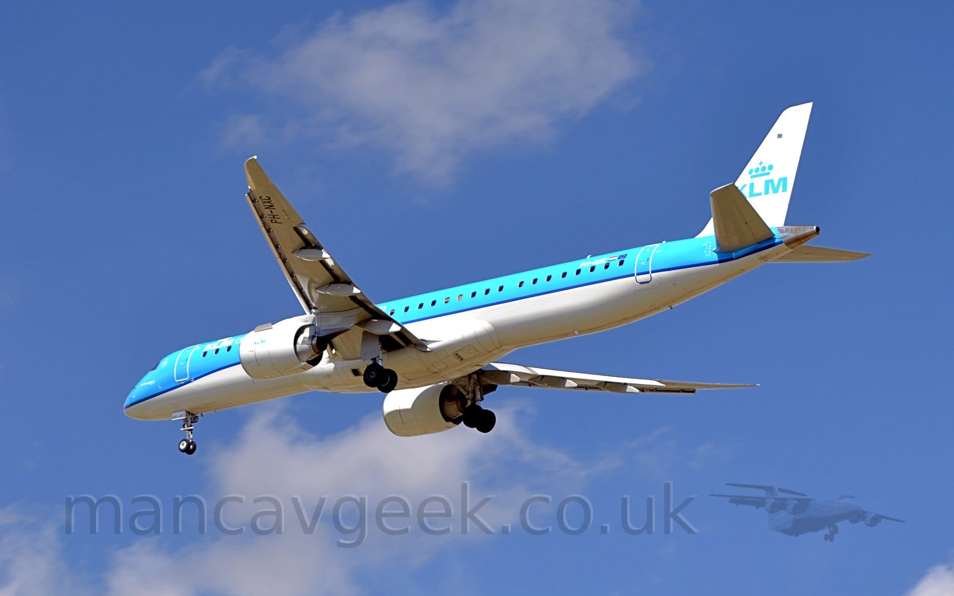 Low side view of a blue and white, twin engined jet airliner flying from right to left at a very low altitude, with undercarriage lowered and flaps extended from the rear of the wings, suggesting it is about to land. The upper fuselage is a pale blue, with a white belly, with dark blue piping separating the 2 colours. There are white letters "KLM" under a stylised white crown on the upper forward fuselage, a logo repeated in blue in a larger form on the white tail, and in smaller form on the sides of the white engine pods under the wings. Pale blue sky with fluffs of white cloud fill the rest of the rain.