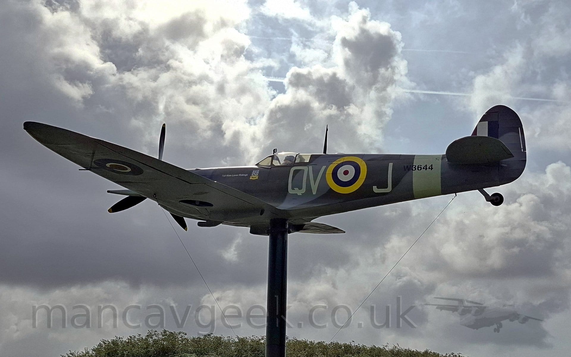 Low side view of a World War 2-era single-engined military fighter pale mounted on a pole in a flying pose. The plane is in a grey and green camouflage scheme, with the cream-coloured leteters "QV-J" either side of a yellow, blue, white, and red roundel on the fuselage. There is a cream band around the rear fuselage, with "W3644" overlaid in black. The underside of the body and wings are a pale creamy white, with a blue, white, and red roundel on the outer panels of the wings. The black pole it sits on seems to rise out of the top of a stand of trees, with dramatically lit grey clouds and blue sky above.