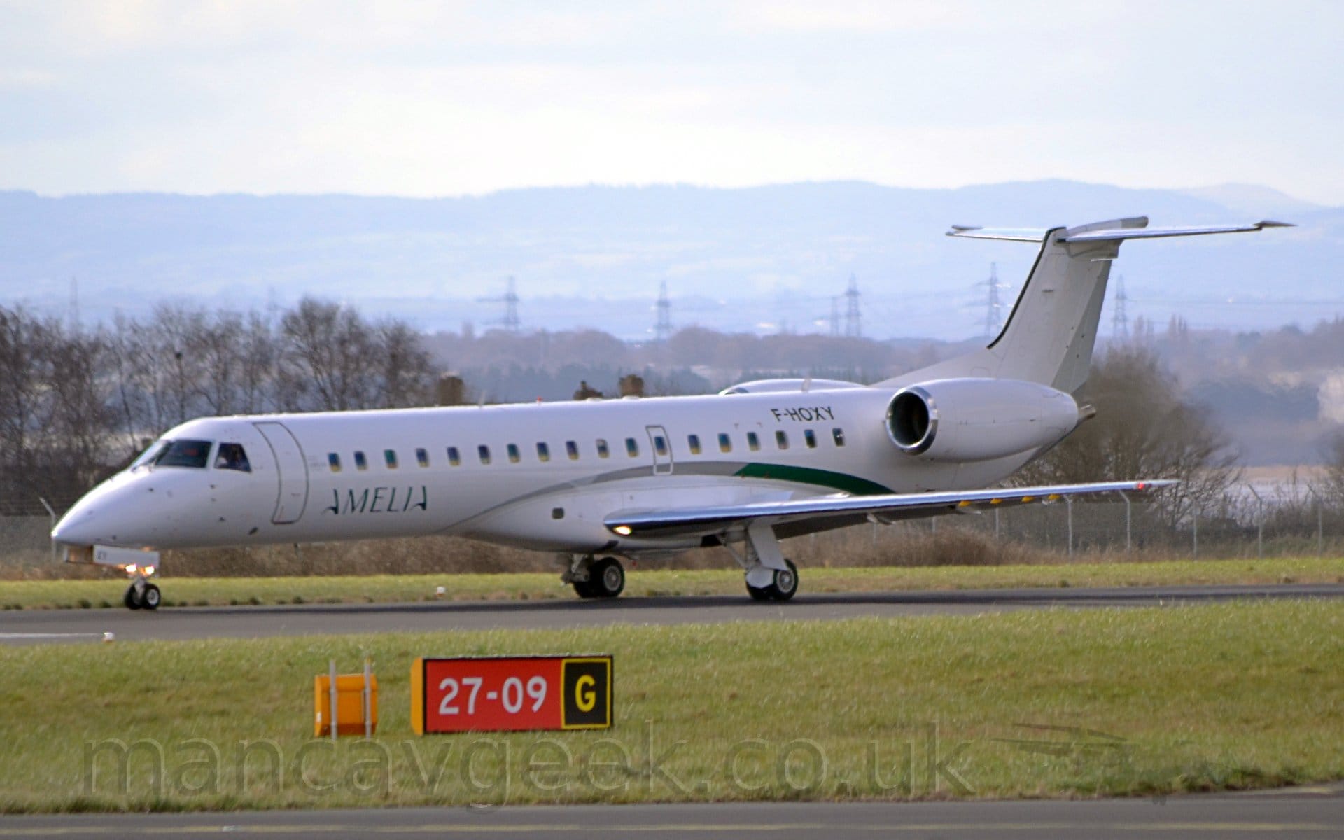 Side view of a twin engined jet airliner taxiing from right to left on a black runway. The plane is almost entirely white, with a grey and green stripe swooping over the wing root. There are green "Amelia" titles on the lower forward fuselage, and the black registration "F-HOXY" on the upper rear. The foreground is filled with green grass, with a red sign with white numbers "27-09"  in the middle and the yellow letter "G" in a black square outlined in yellow in the centre of that grass. Trees fill the background, with hills rising into hazy grey skies in the distance.