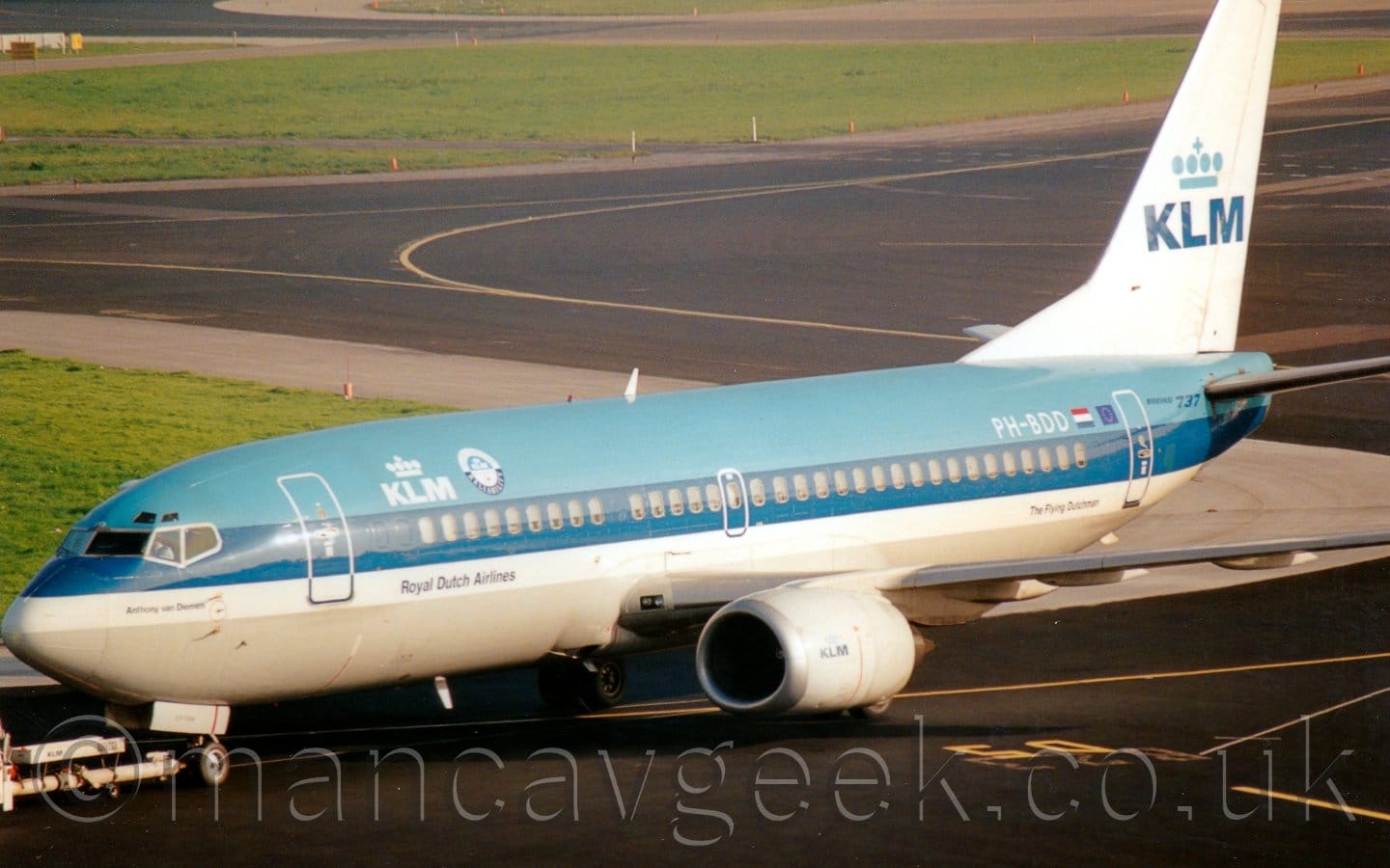 Side view of a pale blue and white, twin engined jet airliner facing to the left but being pushed back to the right by an out-of-frame tug attached to the nose-wheel by a long white pole. The planes upper body is pale blue, with a creamy-grey belly, a thick dark blue stripe running along the body covering the passenger cabin windows, and a thinner white stripe below that. There is white logo on the upper forward fuselage, with white upper-case letters "KLM" under a stylised white crown, a design repeated in a larger form in blue on the white tail. There are small, blue "Royal Dutch Airlines" titles on the white stripe under the main logo. There are additional, small, "KLM" logos on the sides of the white engine pods under the wings. Black taxiways with yellow lines for the planes to follow fill most of the frame, with large strips of grass separating them.