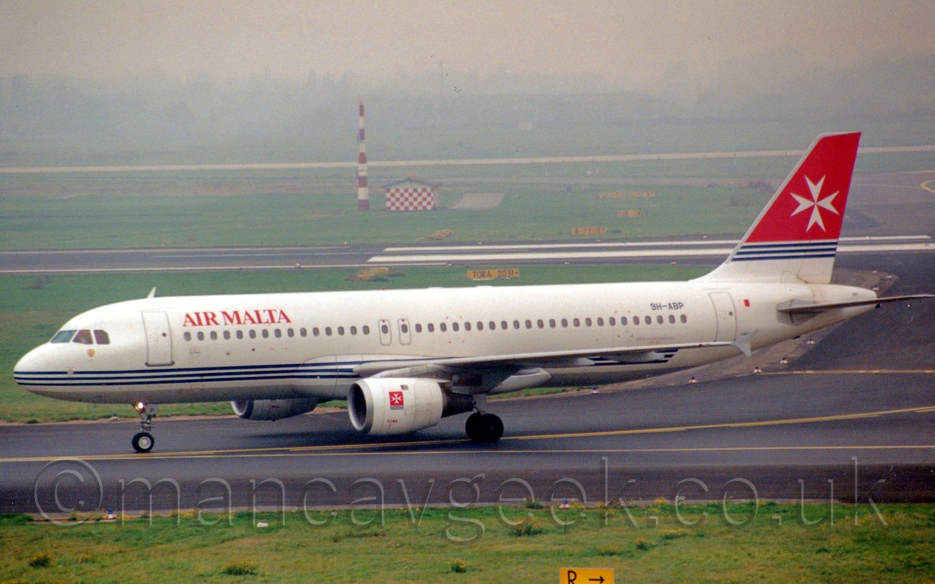 Side view of a white, twin engined jet airliner taxiing from right to left. The plane has a white body with a grey belly, and 3 thin blue stripes running along the lower fuselage, with red "Air Malta titles on the upper forward fuselage. The tail has a red top half, with the bottom being white, separated by 3 thin blue stripes. A large white Maltese Cross is in the middle of the red part of the tail, a design repeated in a square on the side of the white engine pods. Green grass lines the taxiway at the bottom of the frame, and runs off into the distance in the background, slowly vanishing into grey haze.
