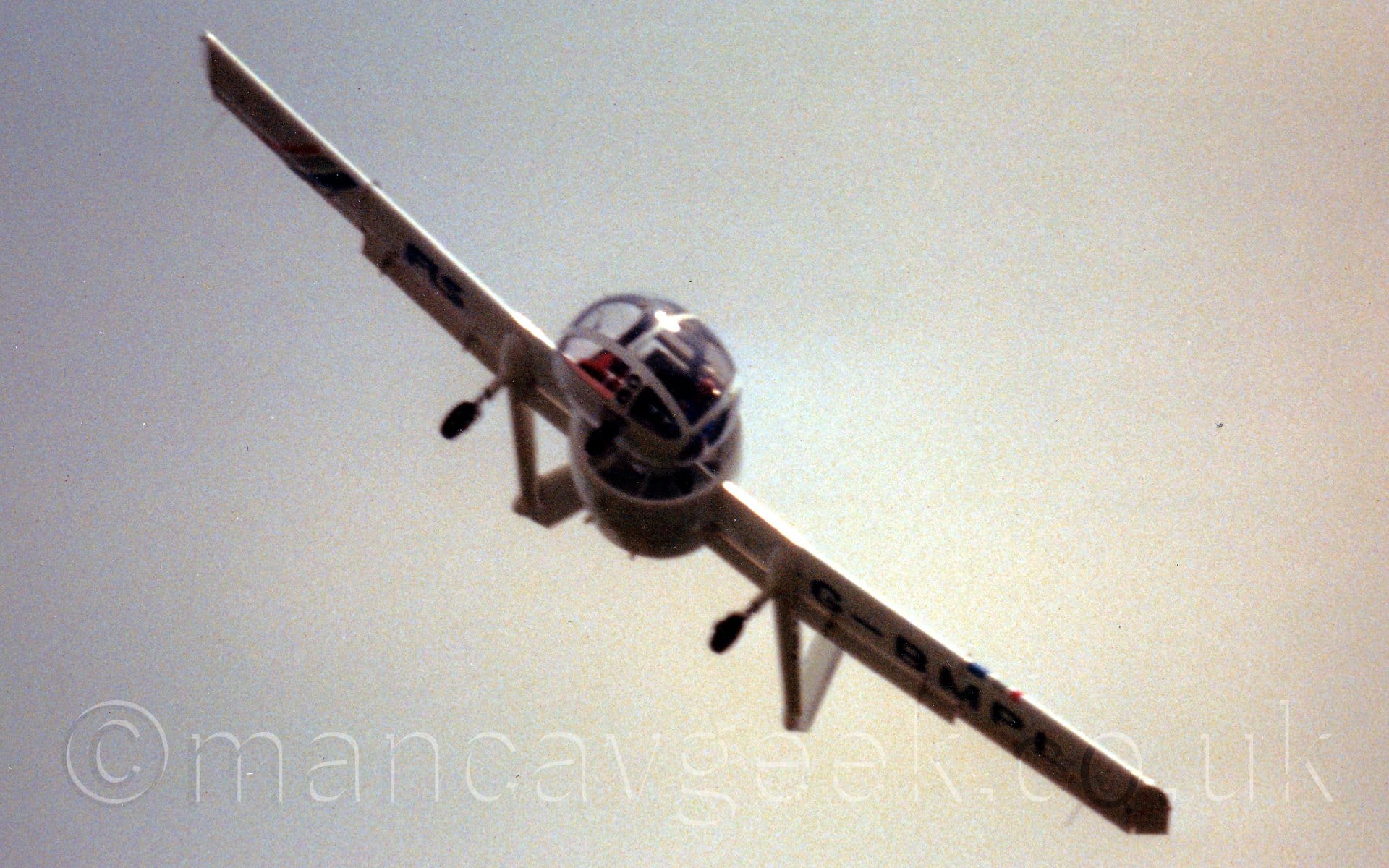 Low, almost head-on view of a white, single engined light aircraft with a bulbous, heavily glazed cockpit canopy in front of a fuselage that is basically a shrouded propellor engine in the middle of a long, straight wing. There are long tail-booms extending part way through each of the wings, with the fixed undercarriage attached to the front of the booms. The plane is heavily tilted to the side, obviously performing a spirited flying display routine. Grey sky fills the rest of the frame.