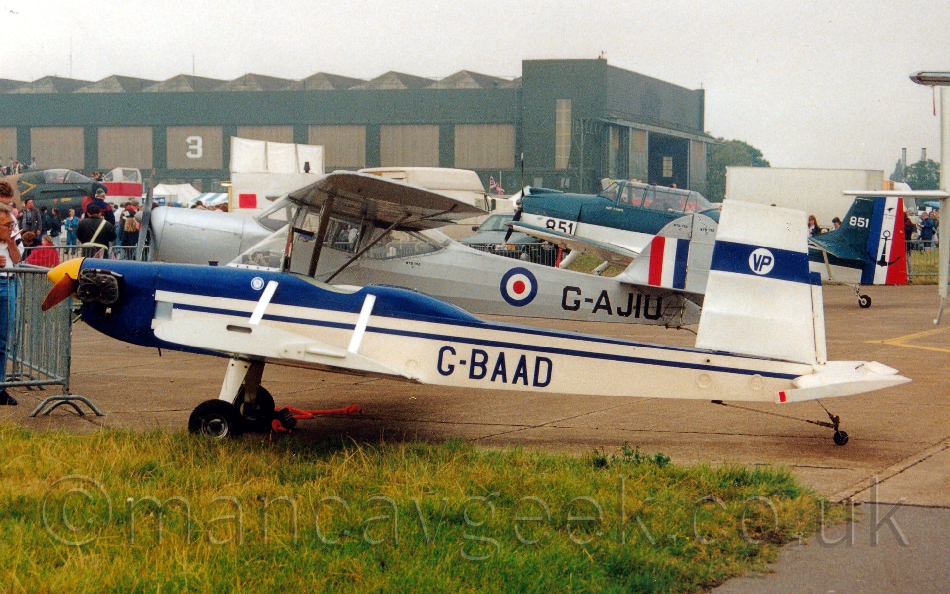 Side view of a cream and blue, long and thin single-engined light aircraft parked facing to the left. A glazed, low windshield is the only concession to protection for the open cockpit and any pilot that may fly this. The plane has dark blue upper surfaces and a cream body, with a white tail with a blue stripe containing a white circle with the letters "VP" in the middle in blue. The registration "G-BAAD" are on the side of the rear fuselage, also in blue. A pair of other single-engined light aircraft, both with military markings, are parked behind this one. Grey metal fencing surrounds these planes, with crowds of people on the other sides, as well as a couple of vehicles. A large green and brown hangar stretches across the frame from the left, under grim grey sky.