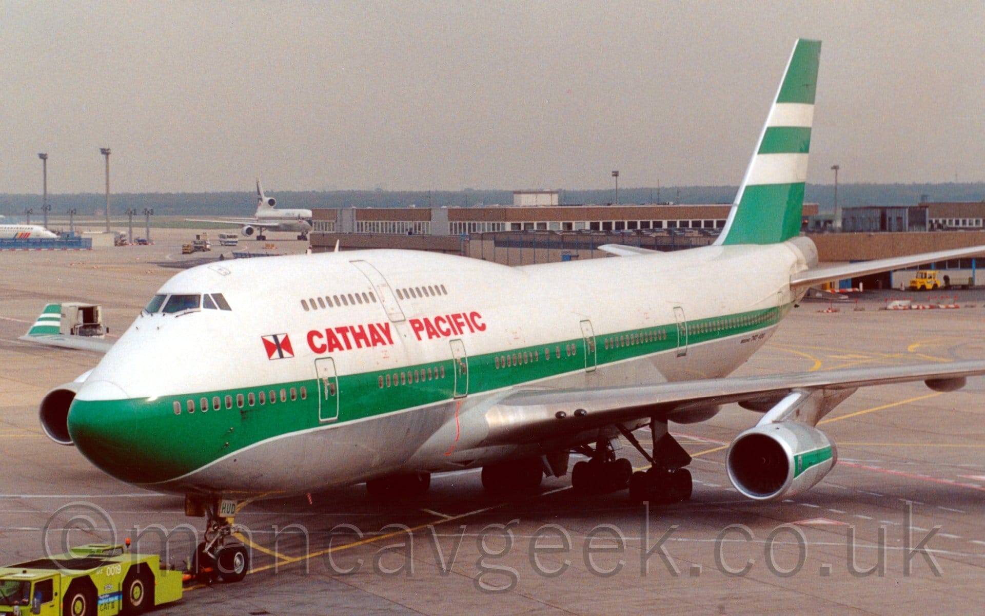 Side view of a very large, white and green, 4 engined jet airliner facing to the left but being pushed back to the right by a lime green tug attached to the main wheel by a long orange pole. The plane is mostly white, with a thick green stripe running along the body, covering the passenger windows on the lower deck, and a grey belly, with white piping. The tail is mostly green, with 2 thick white stripes in the middle. The engine pods under the wings are grey, with a thick green stripe. There are red "Cathay Pacific" titles on the upper forward fuselage, between the upper and lower deck windows. A brown building stretches across the frame from the right in the background, with a white, 3 engined jet airliner just taxiing behind it. Trees line the horizon, under grim grey skies.