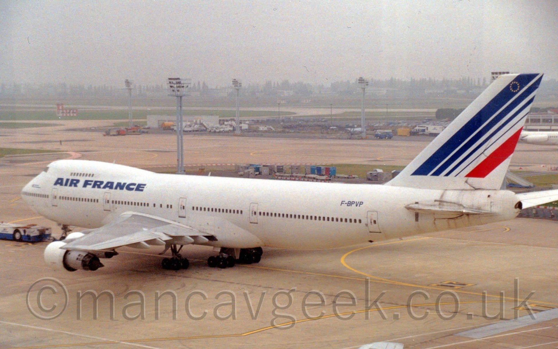 Side view of a very large, white, 4 engined jet airliner being towed from right to left. The plane is almost entirely white, with blue "Air France" titles between the upper and lower decks at the front of the fuselage, and a series of blue diagonal stripes (and a single red stripe) on the tail. The blue registration "F-BPVP" is on the upper rear fuselage, just in front of the tail. A white and blue tug is attached to the nose, pulling the plane across the frame. Tall lighting poles are scattered around the background, with grassed aeras and vehicle parks separating taxiways, slowly vanishing into haze in the distance.