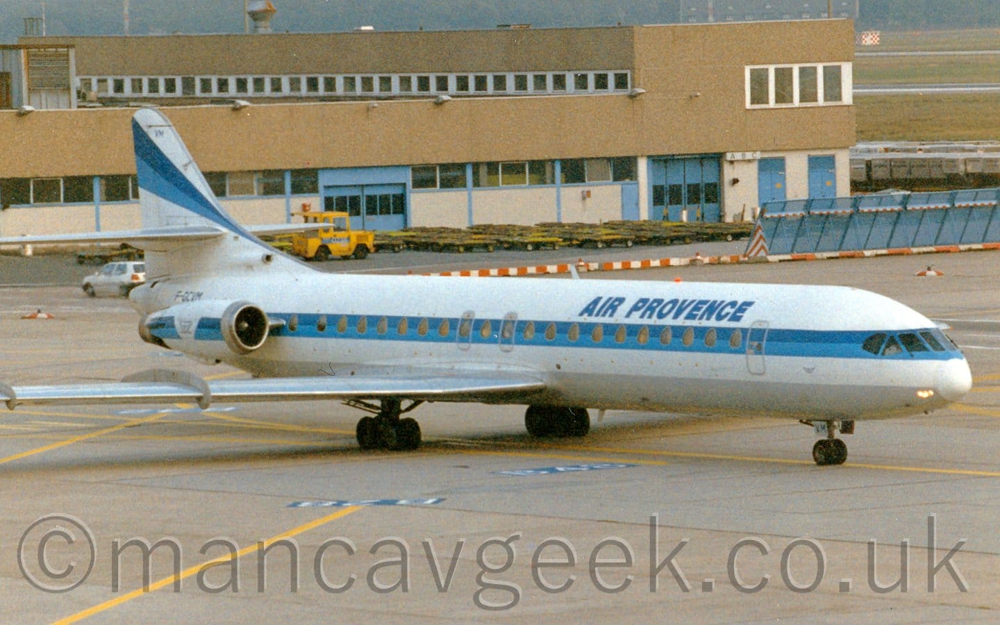 Side view of a white, twin engined jet airliner with the engines mounted on the sides of the rear fuselage, taxiing from left to right. The plane is mostly white, with a dark and light blue stripe running along the body, covering the passenger cabin windows, a grey belly. There are dark blue "Air Provence" titles on the upper forward fuselage, and the blue registration "F-GCVM" on the upper rear, under the tail. Grey concrete apron marked with yellow lines fills the foreground, with a sandy brown building stretching across the background from the left.