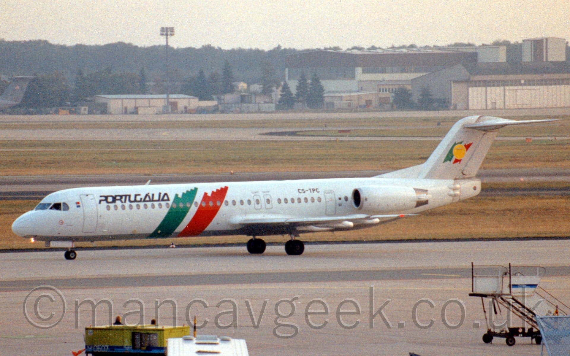 Side view of a white, twin engined jet airliner with the engines mounted on the sides of the rear fuselage, taxiing from right to left. The plane is mostly white, with diagonal green and red bands running from the top of the body, down and forward under the belly. There are black "Portugalia" titles on the upper forward fuselage, just aft of the forward door. The tail is mostly grey, with a smaller version of the green and red bands body bands overlaid with a yellow circle. Grey concrete apron dills the foreground, with grass and taxiways in the background lead up to cream-coloured hangars and other buildings in the distance, under bright but hazy sky.