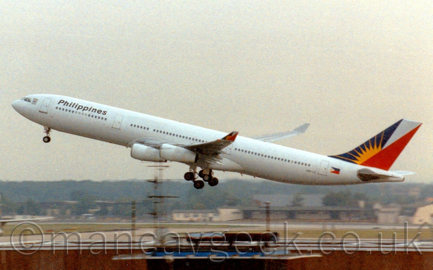 Slightly grainy side view of a white, 4 engined jet airliner flying from right to left at a very low altitude, with undercarriage lowered, flaps extended from the rear of the wings, and the nose raised sharply, suggesting it has just taken off. The planes body is all white, with black "Philippines" titles on the upper forward fuselage, and blue, white, and red triangles on the tail overlaid with a yellow sun-burst. A similar logo appears on the up-turned wing-tips. The top of a brown building stretches across the bottom of the frame, with cream-coloured buildings in the distance, with trees beyond that, under grim grey sky.