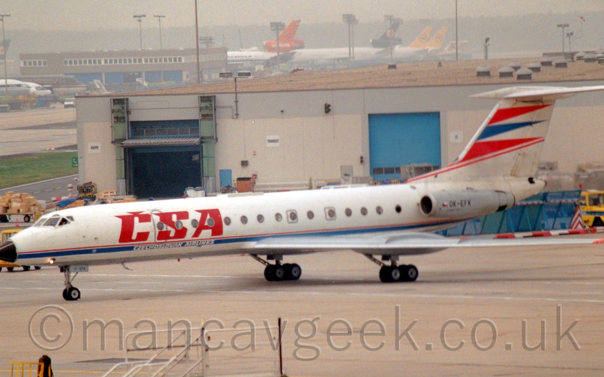 Side view of a white, twin engined jet airliner with the engines mounted on the sides of the rear fuselage, taxiing from right to left. The plane is mostly white, with large red "CSA" titles on the sides of the forward fuselage, with white "Czechslovak Airlines" text just below on a thin blue stripe running along the body. A pair of red triangles separated by a blue triangle adorn the leading edge of the tail, with red piping underneath. The engine pods on the rear fuselage are grey, with the registration "OK-EFK" in black. A white building with a flat, sandy brown roof and blue roller doors stretches across most of the frame from the right, with the tails of other jet airliners next to a brown building in the distance.