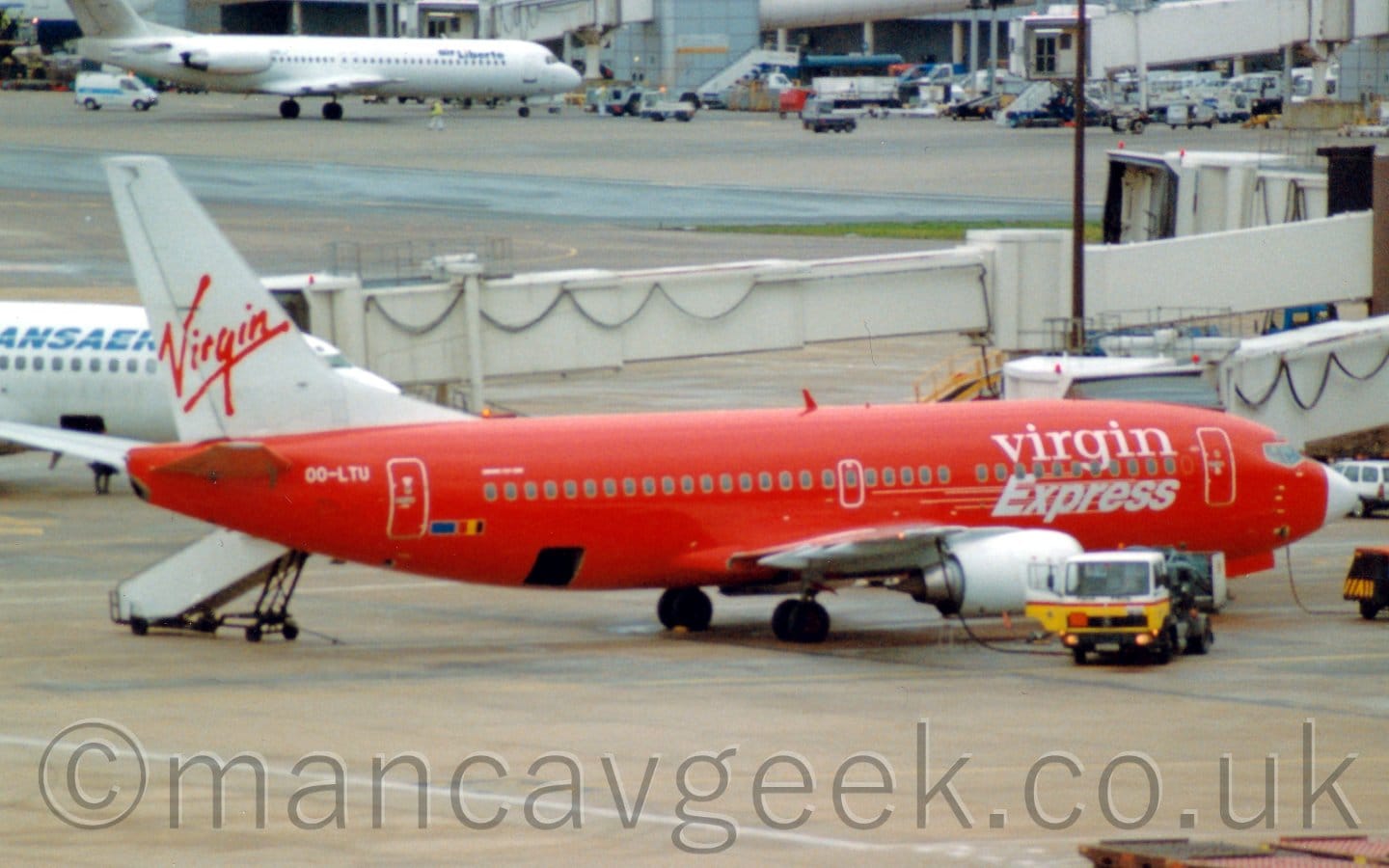 Side view of a red and white, twin engined jet airliner parked facing to the right. The plane has a red body, with white "Virgin Express" titles on the forward fuselage, and a white nose cone. The tail is white, with red, seemingly hand-written "Virgin" titles. A yellow and white fuelling truck is parked next to the white engine under the right wing, with a fuel hose running into one of the wings internal fuel tanks. Grey concrete apron fills the foreground, long, cream-coloured airbridges stretching across the frame in the background from the right, one attached to this plane, another to a white plane behind it on the left. More concrete apron leads off to a grey terminal building in the distance at the top of the frame, with a white jet airliner parked next to it.