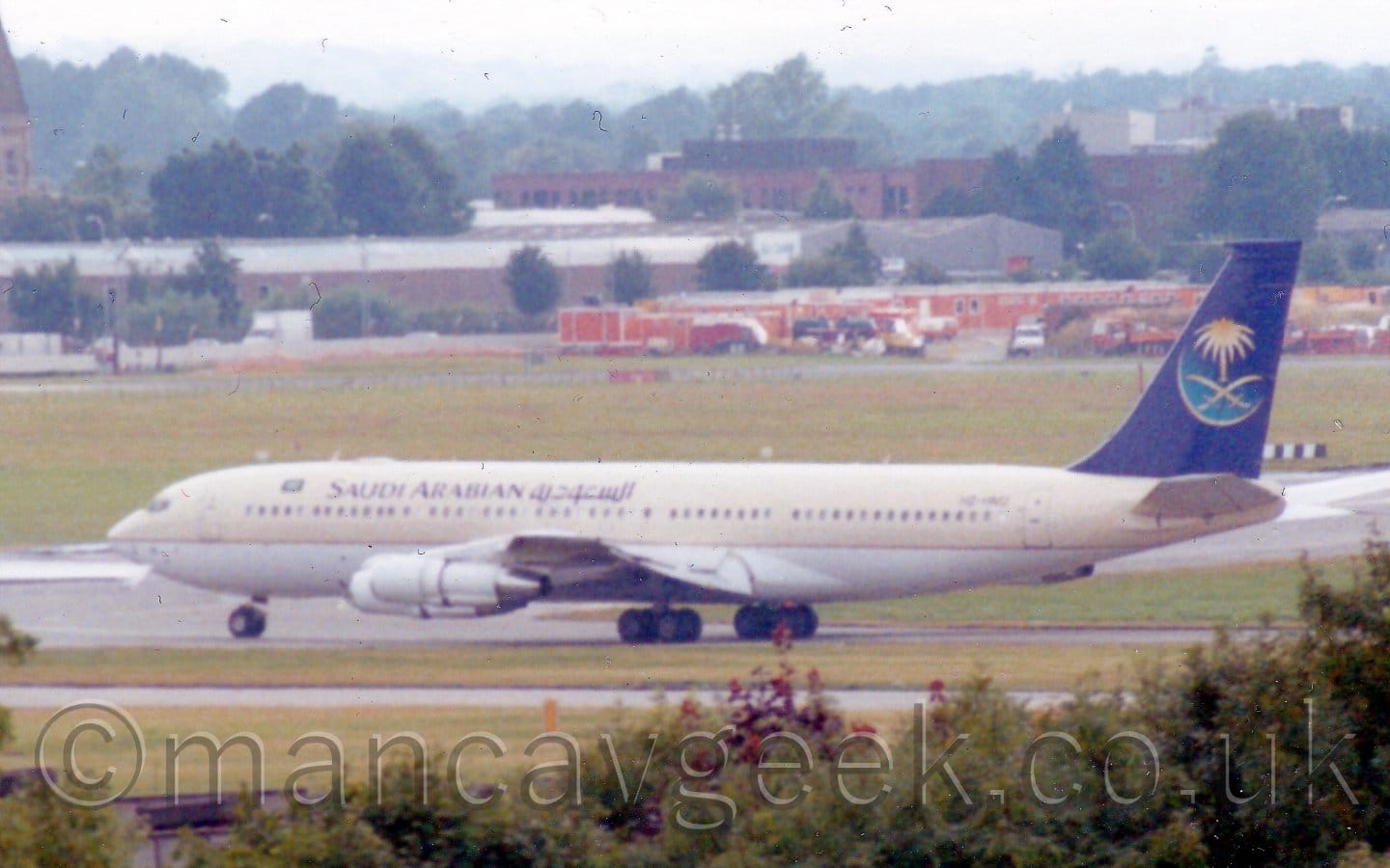 Slightly grainy and blurry side view of a creamy-coloured, 4 engined jet airliner taxiing from right to left, The plane has creamy upper surfaces, and a white belly. There are dark blue "Saudi Arabian" titles on the upper forward fuselage. The tail is the same dark blue, with a pale blue crescent moon, crossed golden sabres, and golden palm tree. Trees and bushes line the bottom of the image, with grass leading up to buildings and trees in the distance.