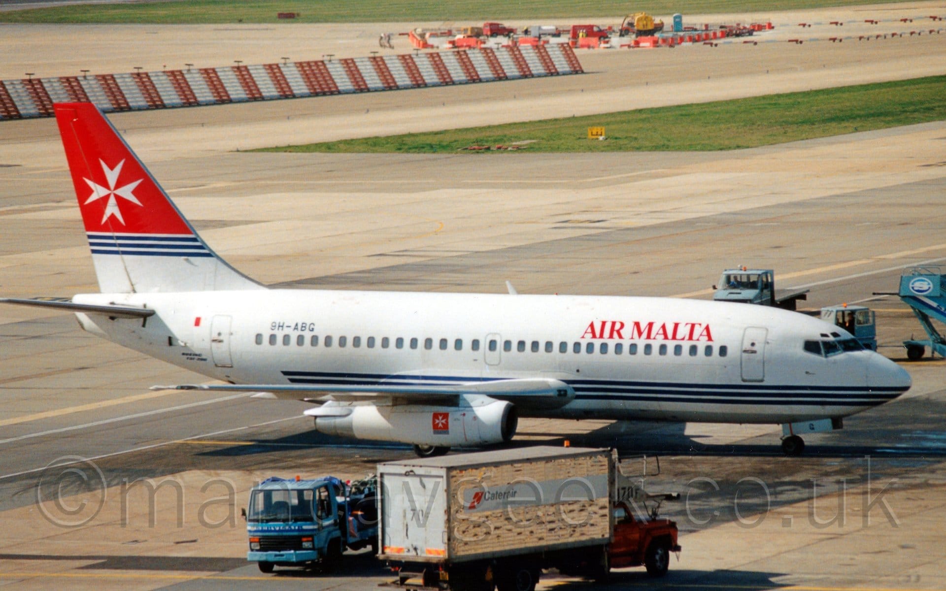 Side view of a white, twin engined jet airliner taxiing from left to right. The plane has 3 thin blue stripes running along the body, on a level with the wing root, and red "Air Malta" titles on the upper forward fuselage. The registration "9H-ABG" is on the upper rear fuselage in black. The bottom of the tail is white, with3 thin blue stripes at the top of that section, while the top is red with a white Maltese Cross (4 arrow-heads with the tips meeting in the middle.) A smaller version of the same logo (white Maltese Cross on a red background over 3 blue lines) is on the side of the engine pods. Grey concrete apron fills most of the frame, with a white van and a blue fuel truck in the middle of the foreground. More blue trucks and a blue set of mobile air stairs are in the background on the left. A strip of green grass separates 2 taxiways, with a red and white striped metal fence and a small building site separating them from another in the distance.