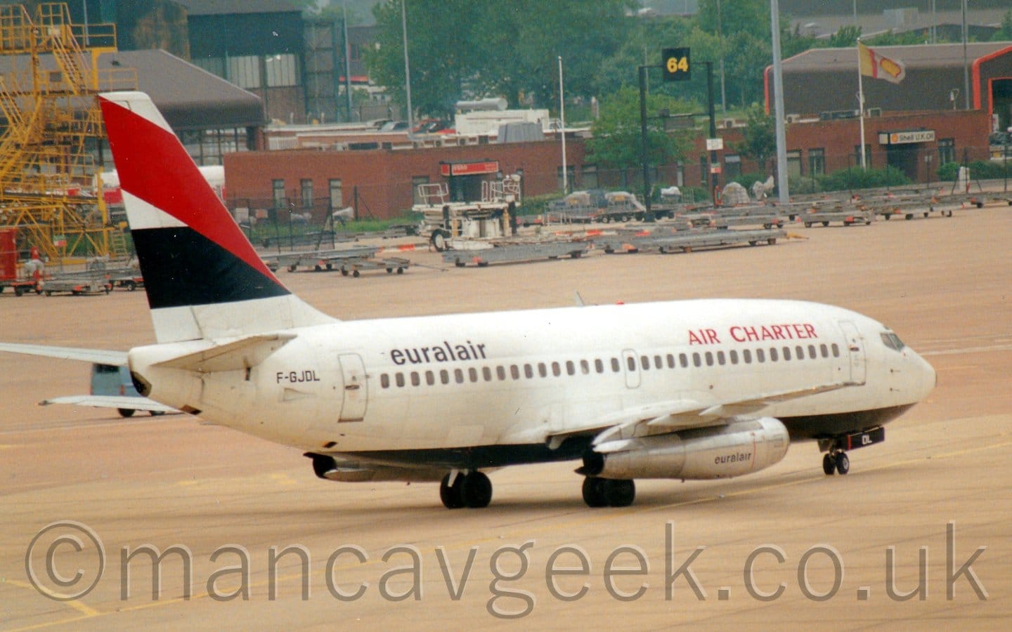Side view of a white, twin engined jet airliner taxiing from left to right. The plane is mostly white, with a black belly, and red "Air Charter" titles on the upper forward fuselage, and black "EuralAir" titles on the upper rear. There is a thick black stripe across the tail, overlaid with a thick diagonal red band. The black registration "F-GJDL" is on the rear fuselage, under the tail. Grey concrete apron fills most of the foreground and part of the background, with rows of grey freight trolleys lined up in the distance, with brown brick buildings and trees beyond that.