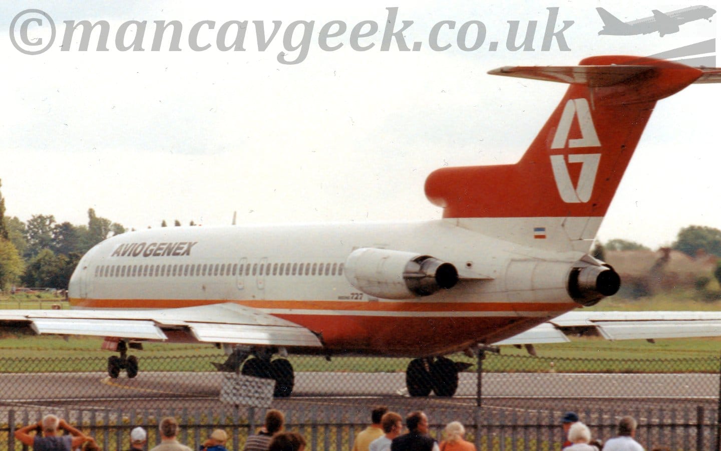 Side view of a 3 engined jet airliner with the engines mounted on the rear of the fuselage, taxiing from right to left as it turns from a taxiway on to a runay. The plane is mostly white, with an orange stripe running along the lower body, and a red belly. There are small, black "Aviogenex" titles on the upper forward fuselage. The tail is red, with a stylised white letter "A", with a reflection below. A black chain-link fence stretches across the bottom of the frame, with people standing around, some looking at the plane. Green grass on the far side of the runway leads off to trees in the distance, under grey skies.