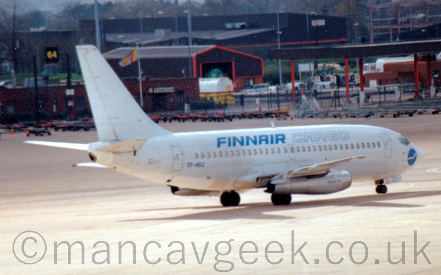 Side view of a white, twin engined airliner taxiing from left to right. The plane is almost entirely white, with blue "FinnAir" titles on the upper rear fuselage, and white "Cargo" titles outlined in blue on the upper forward fuselage. There is a blue circle with a stylised letter "f" with a long tail in the middle on the nose under the cockpit. The registration "TF-ABJ" is on the lower rear fuselage in blue. Grey concrete apron fills the foreground, and leading up to brown buildings with red trim in the background, with trees behind them in the distance.
