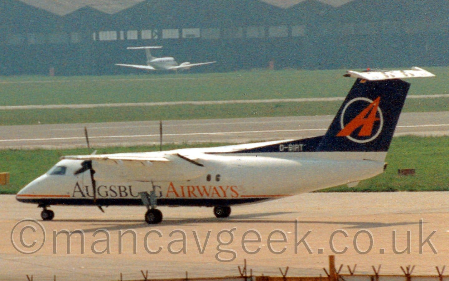 Side view of a high-winged, twin propellor-engined airliner taxiing from right to left. The plane has a white body, with a blue belly, and a red pinstripe overlaid with blue and red "Augsburg Airways" titles. The tail is dark blue, with a red upper-case letter "A" leaning to the right, with the white outline of a circle behind it. Grey concrete apron fills the foreground, with grass either side of a black runway in the background leading up to a wide black hangar that stretches across the frame, with a white BizProp parked in front.
