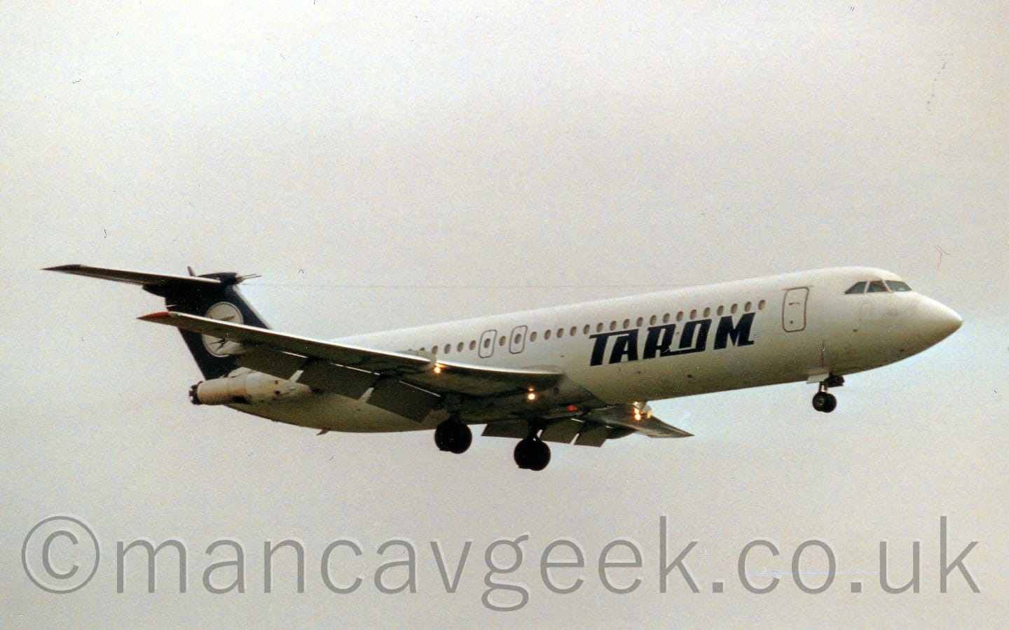 Side view of a white, twin engined jet airliner with the engines mounted on the sides of the rear fuselage, flying from left to right at a very low altitude, with undercarriage lowered and flaps extended from behind the wings, suggesting it is about to land. There are dark blue "Tarom" on the lower forward fuselage. The tail is dark blue, with a white circle containing a dark blue flying bird. Several bright lights are shining forward from under the wings. Flat grey sky fills the rest of the frame.