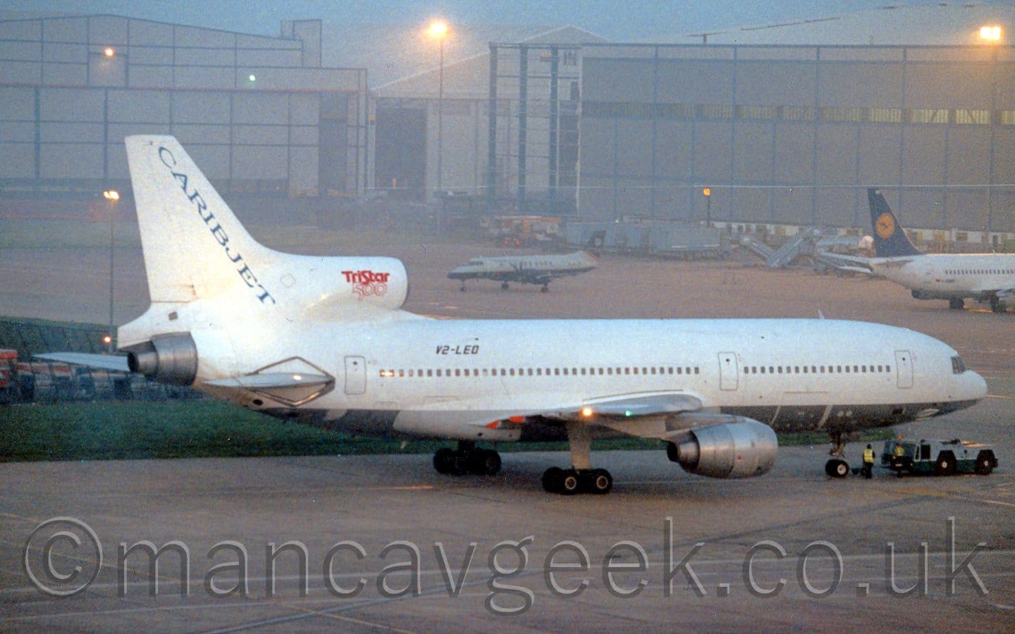 Side view of a white, 3 engined jet airliner facing the right but being pushed back to the left by a low-slung blue and grey tug attached to the nosewheel. The plane has a grey belly, with blue "CaribJet" titles running diagonally down the leading edge of the tail. The registration V2-LEO is on the upper rear fuselage in black, and there are red "TriStar 500" on the centre engine intake. 2 men in hi-viz jackets are standing between the plane's nosewheel and the tug. Grey concrete apron fills the foreground, as well as a large chunk of the background, partly separated by a grassed are on the left of the frame. A pair of large, grey hangars dominate the background, with a pair of other planes parked in front - a grey and almost black twin propellor-engined airliner on the left, and a white twin engined jet airliner with a dark blue and yellow tail on the right. It is obviously dusk, the whole scene being illuminated by bright lights, giving the whole image a slight yellow cast., with grey-blue sky visible between the hangars.